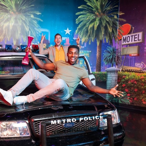 Two smiling men are posing on a "Metro Police" car, one holding a large drink, with a neon motel sign and palm trees in the background.