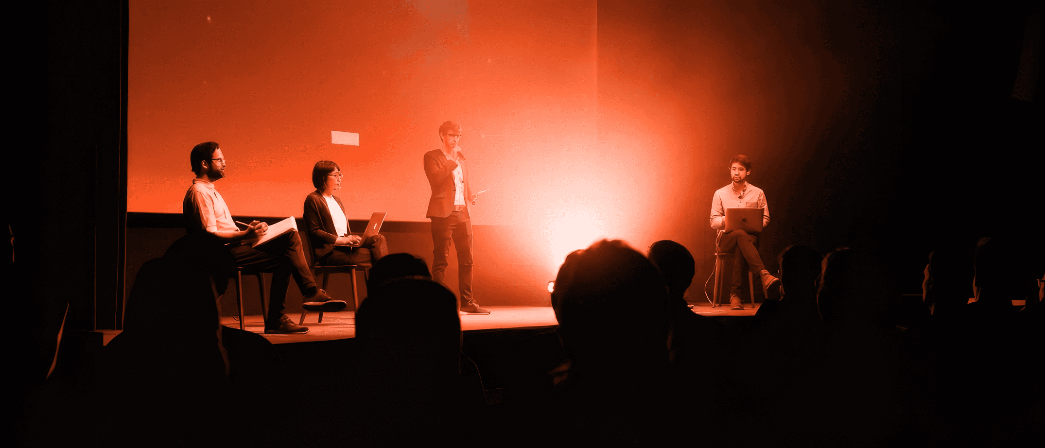 Silhouetted figures on stage against a glowing orange background, performing in a dimly lit venue.