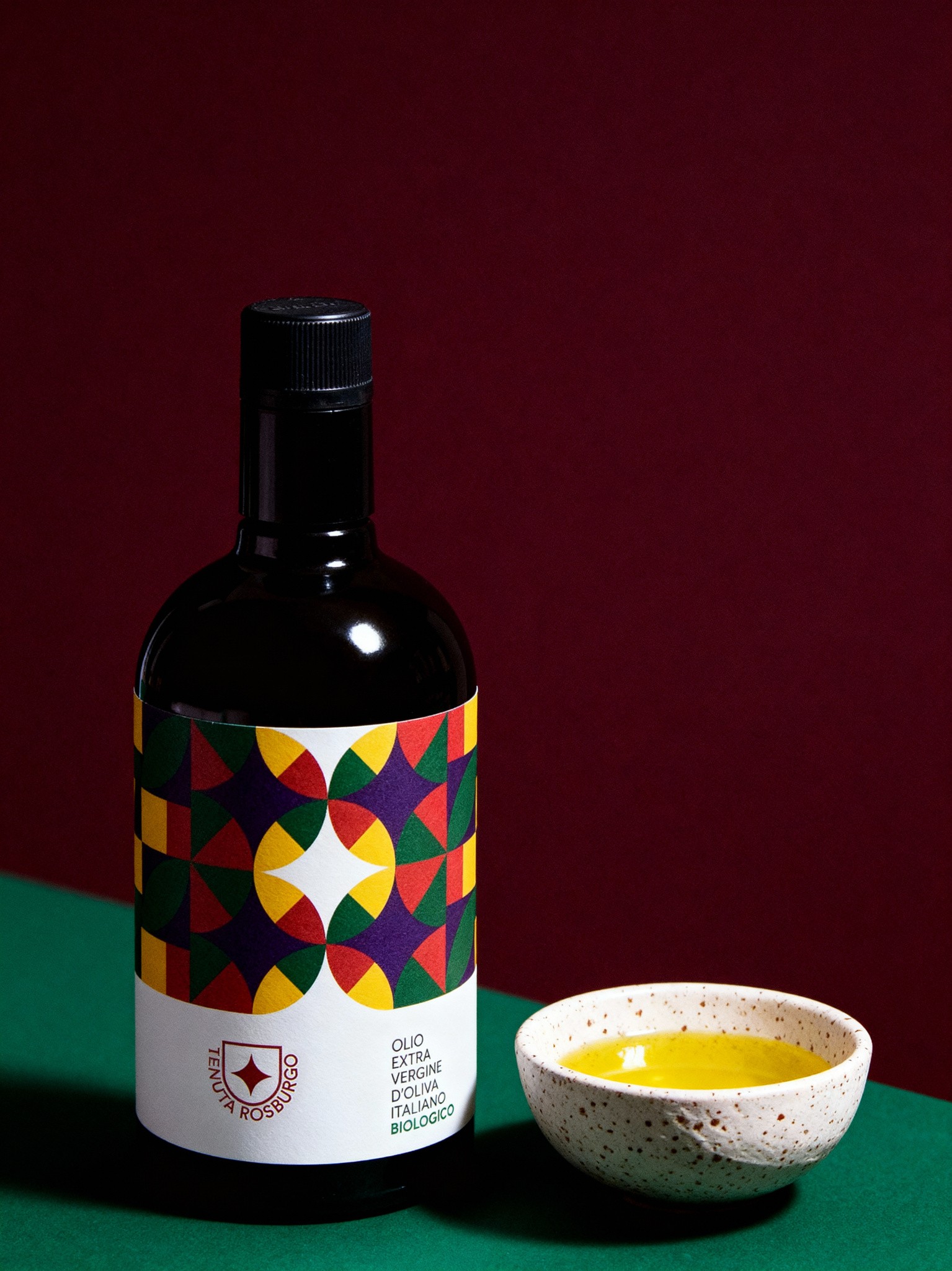 AI product image of Tenuta Rosburgo extra-virgin olive oil bottle with colorful geometric label beside a ceramic bowl of oil, created by Myrovia Labs. AI creative direction for gourmet and lifestyle brands.