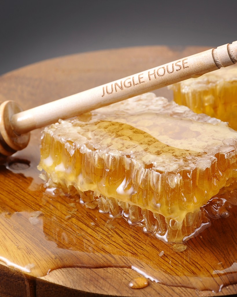 Close-up of honeycomb pieces on a wooden platter with honey dripping, accompanied by a wooden honey dipper engraved with 'Jungle House'.