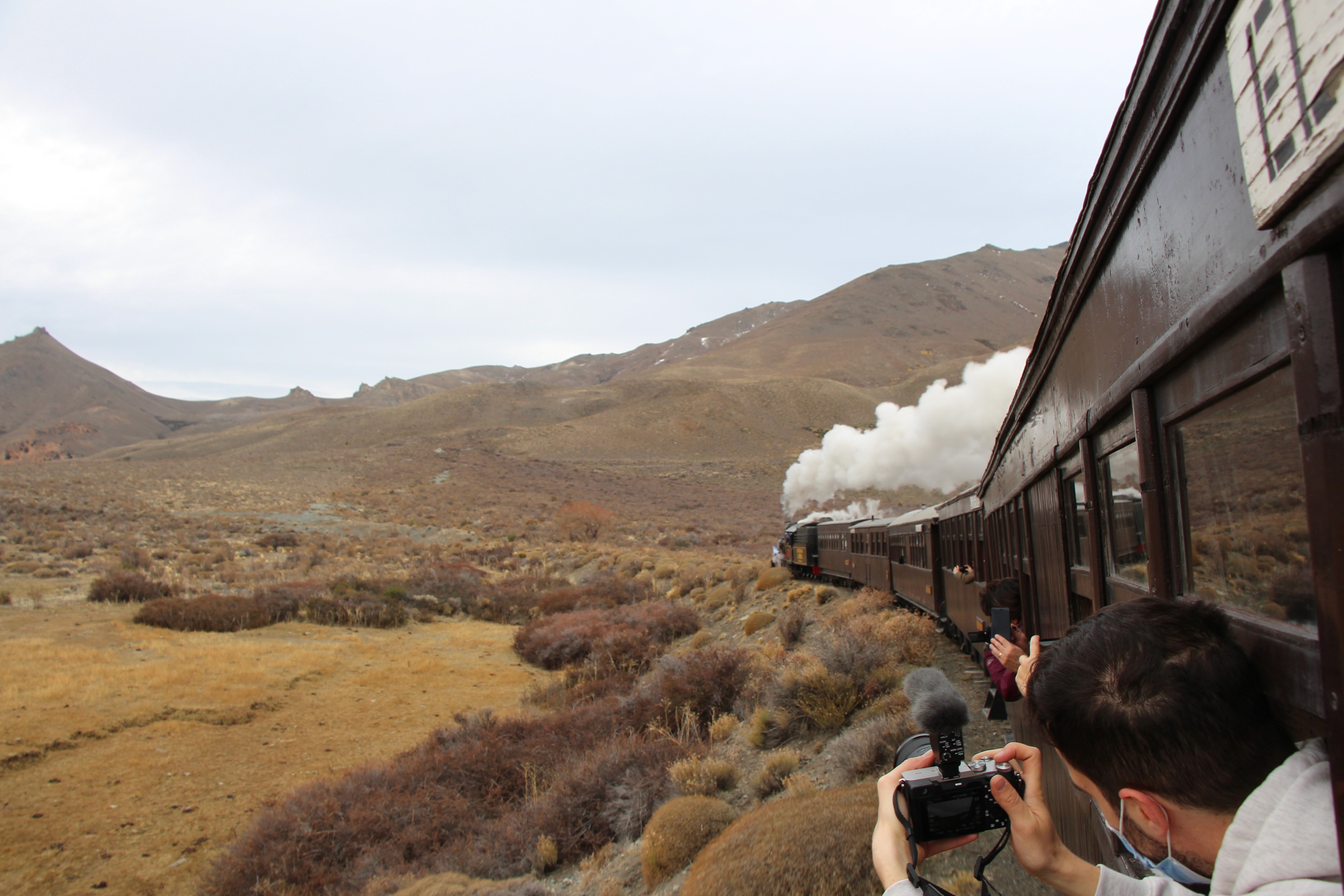 A passenger leans out of a vintage train winding through a dry, mountainous landscape in Patagonia, capturing the moment with cameras as white steam billows from the locomotive ahead.