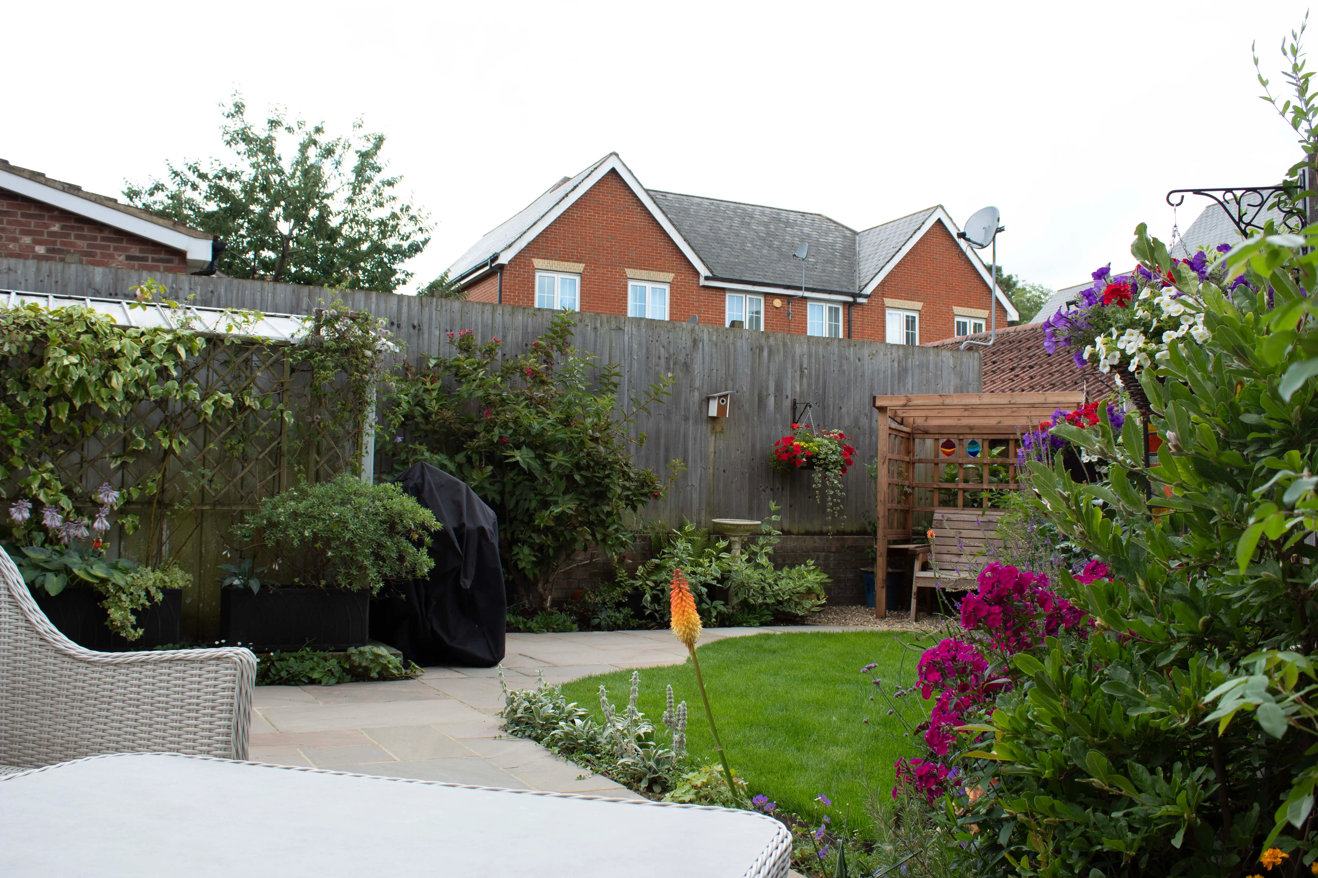 A cozy garden with greenery and flowers, featuring a pathway and houses in the background under a cloudy sky.