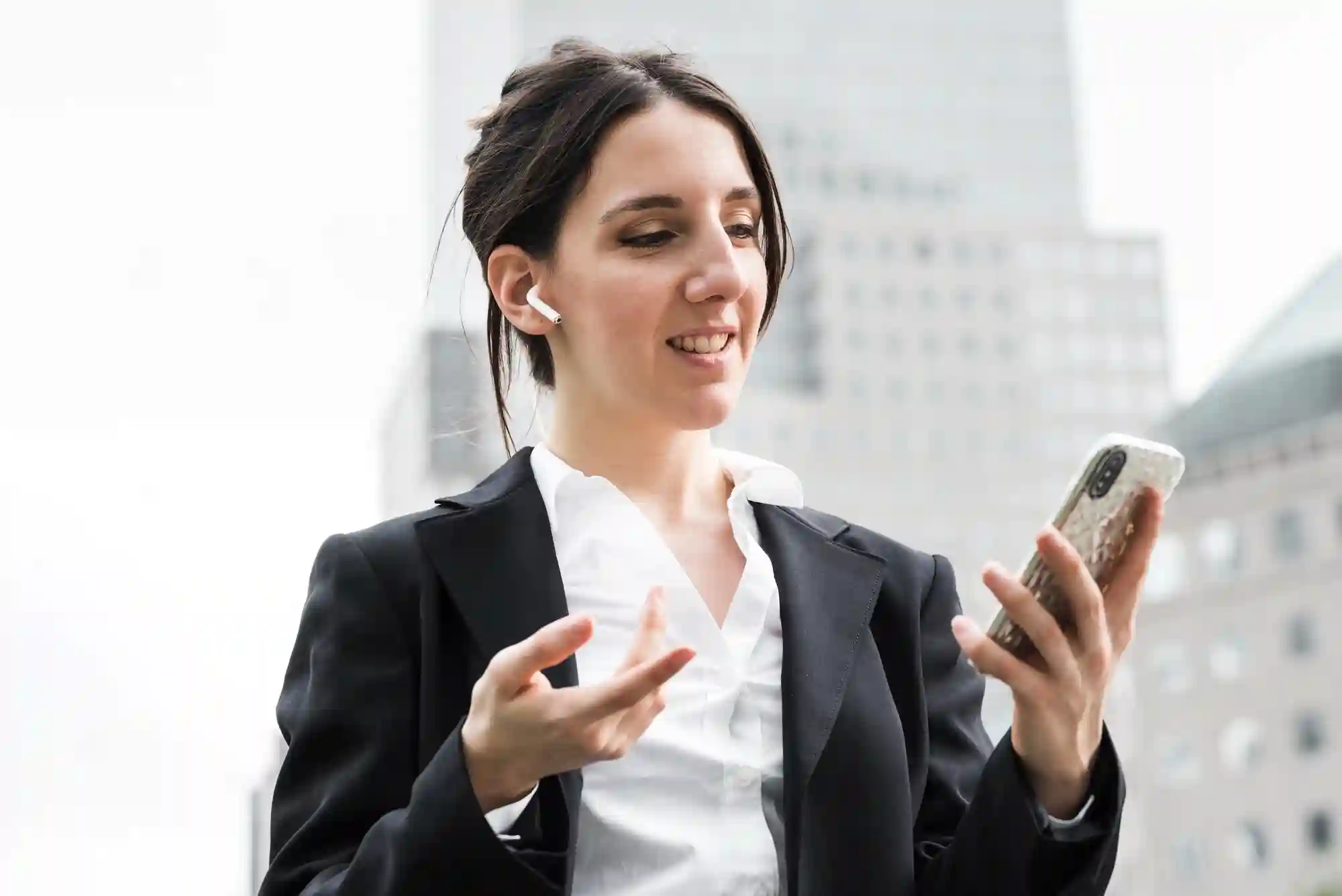 Businesswoman wearing earbuds and talking on a smartphone in a city street, representing remote professional communication.