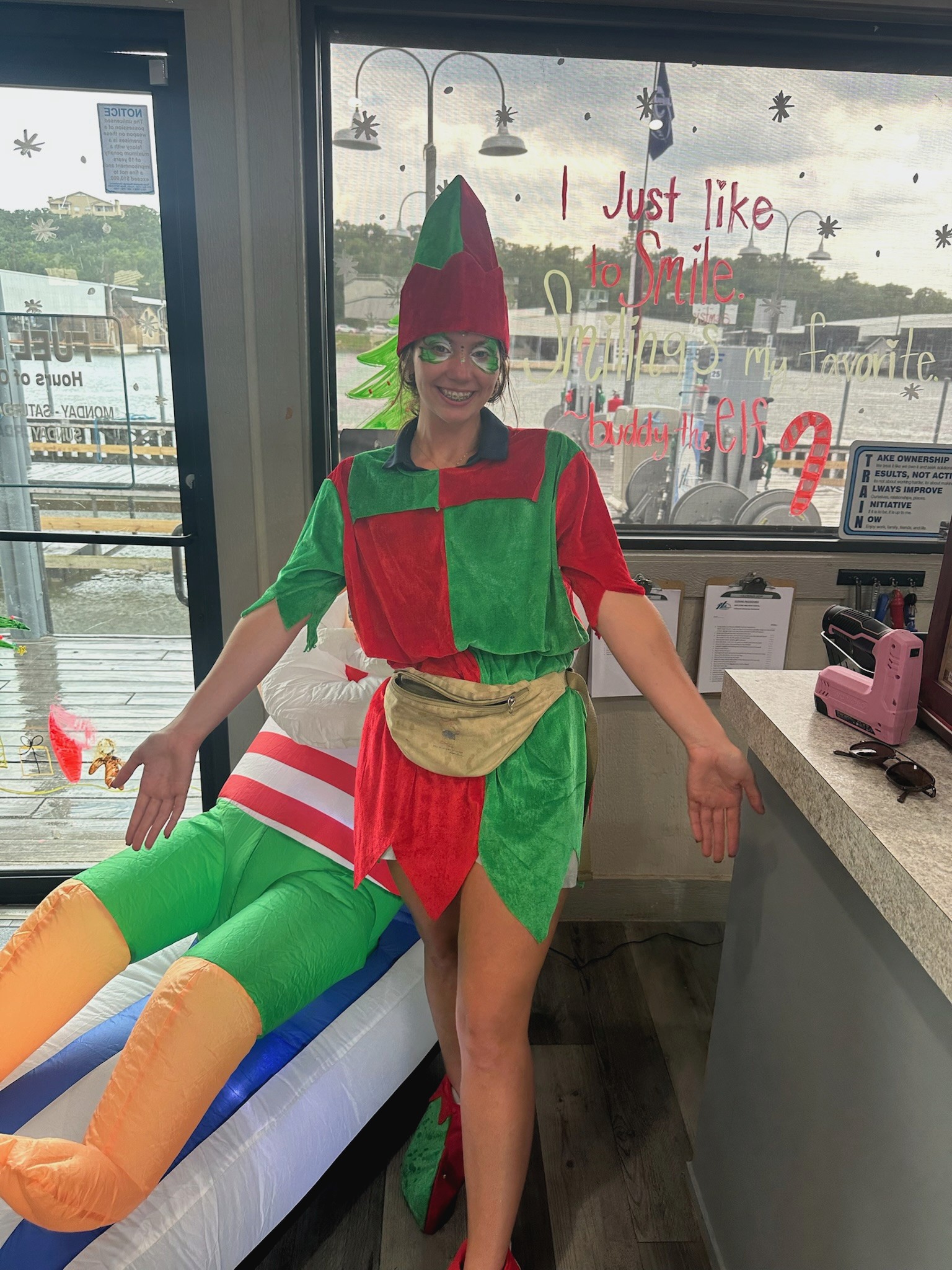 A person dressed as a festive elf in a colorful red and green costume with a pointed hat stands inside a decorated room, next to a large inflatable figure, with a window showcasing a river view in the background; holiday decor includes handwritten window messages and hanging ornaments, creating a cheerful atmosphere.