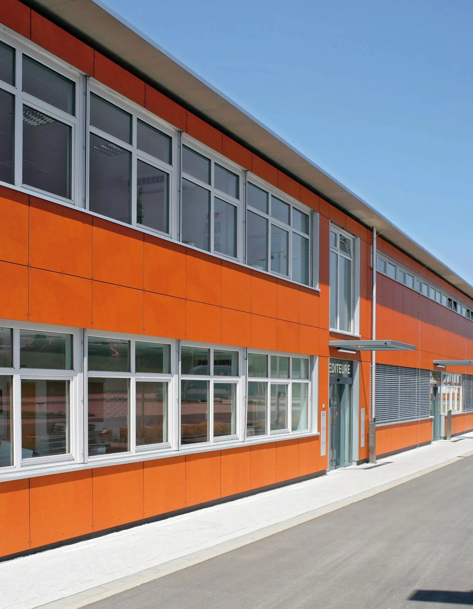 Moderne orange Gebäudezeile mit großen Fenstern und einer Fassade aus hellen Platten, an einem sonnigen Tag.
