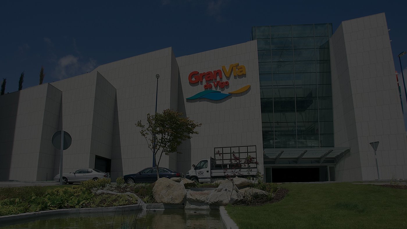 Moderno centro comercial con una gran fachada de vidrio y un colorido cartel "Gran Vía de Vigo". Coches y vegetación en primer plano, cielo azul claro arriba.