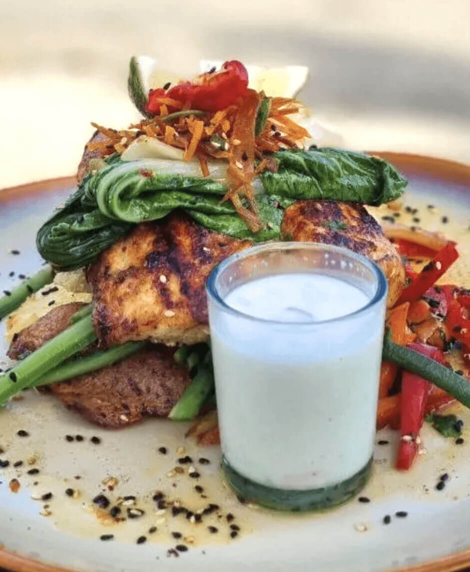 A vibrant dish with grilled fish, bok choy, roasted potatoes, red peppers, and a side of creamy coconut sauce.