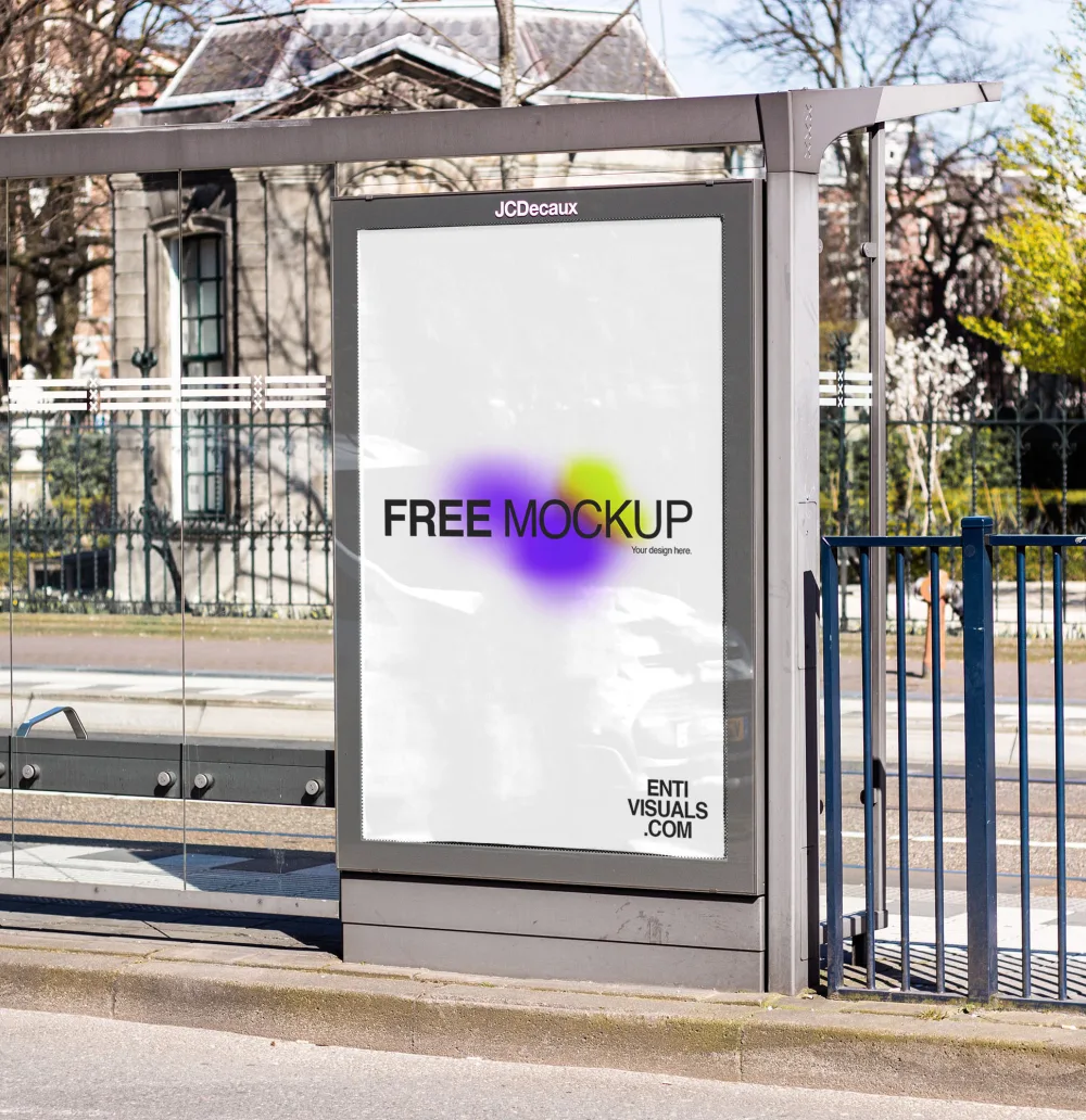 Vertical advertising panel with a blank white poster placed at a modern glass bus stop. The setting includes a sunny urban street, railings, and a residential building in the background.