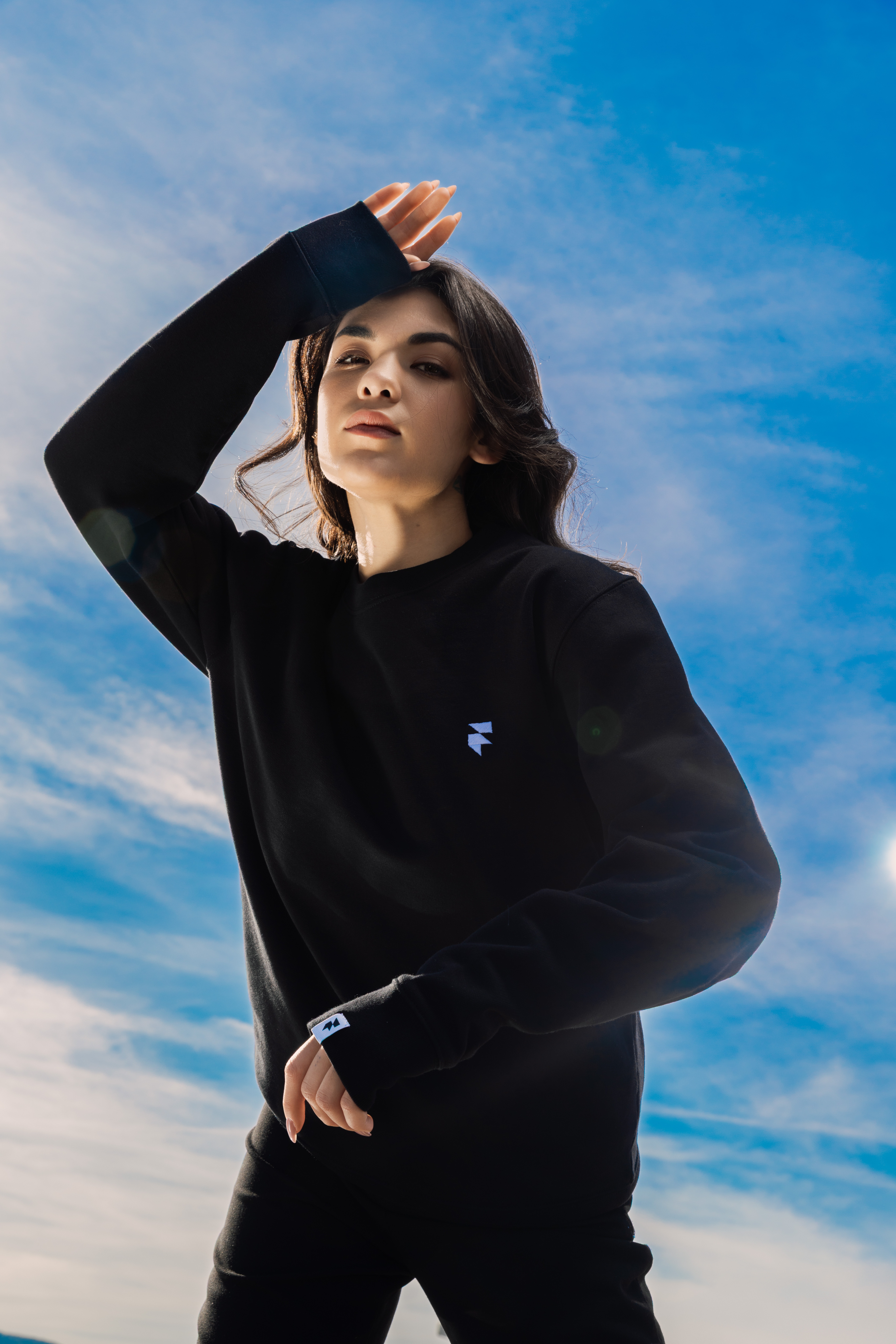 A person with long brown hair wears a black sweater and raises their arm against a vibrant blue sky with wispy clouds.