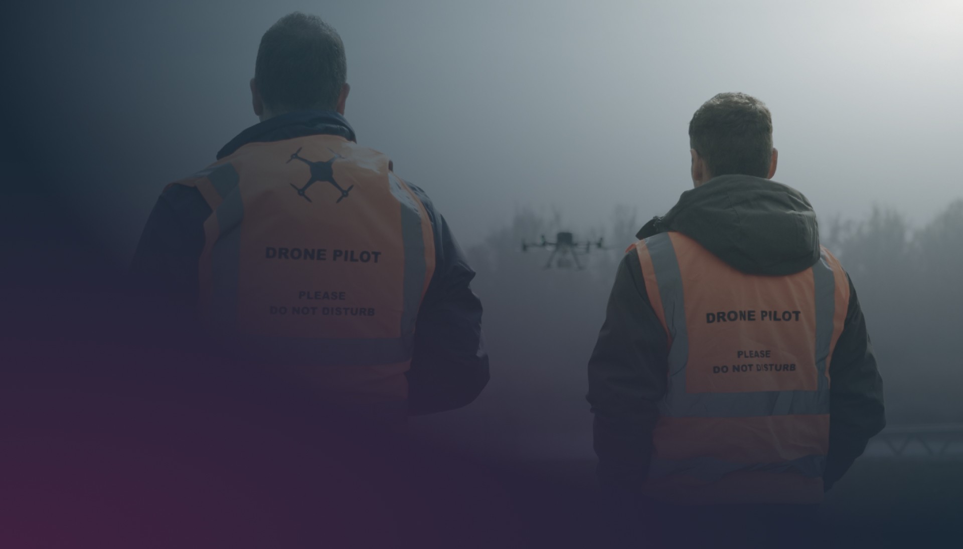 Two drone pilots in orange high-visibility vests watching a drone fly in a misty outdoor environment