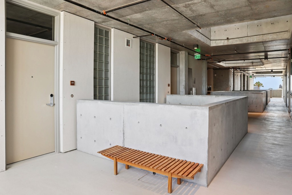 Modern, minimalist corridor with exposed concrete walls and ceiling. A wooden bench sits on the light floor beside a door. Natural light creates a calm ambiance.