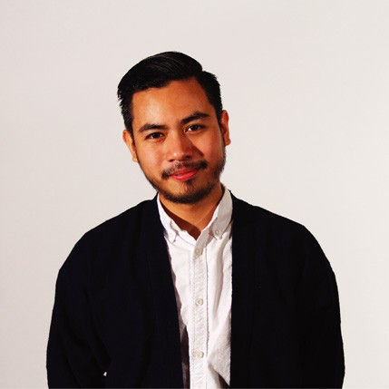 The image shows a man with dark hair and a beard, wearing a black cardigan over a white button-down shirt. He is positioned against a plain off-white or light gray background and is looking directly at the camera with a neutral expression. His hair is styled in a neat, combed-back manner.