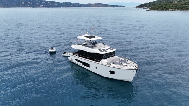 Aerial starboard bow quarter view of the Cranchi T55 Eco Trawler at anchor in a sheltered Mediterranean anchorage, showing the full hull profile with white topsides, large oval hull window, foredeck solar panels, black superstructure with panoramic glazing, flybridge solar array, and a small tender astern against a wooded island backdrop
