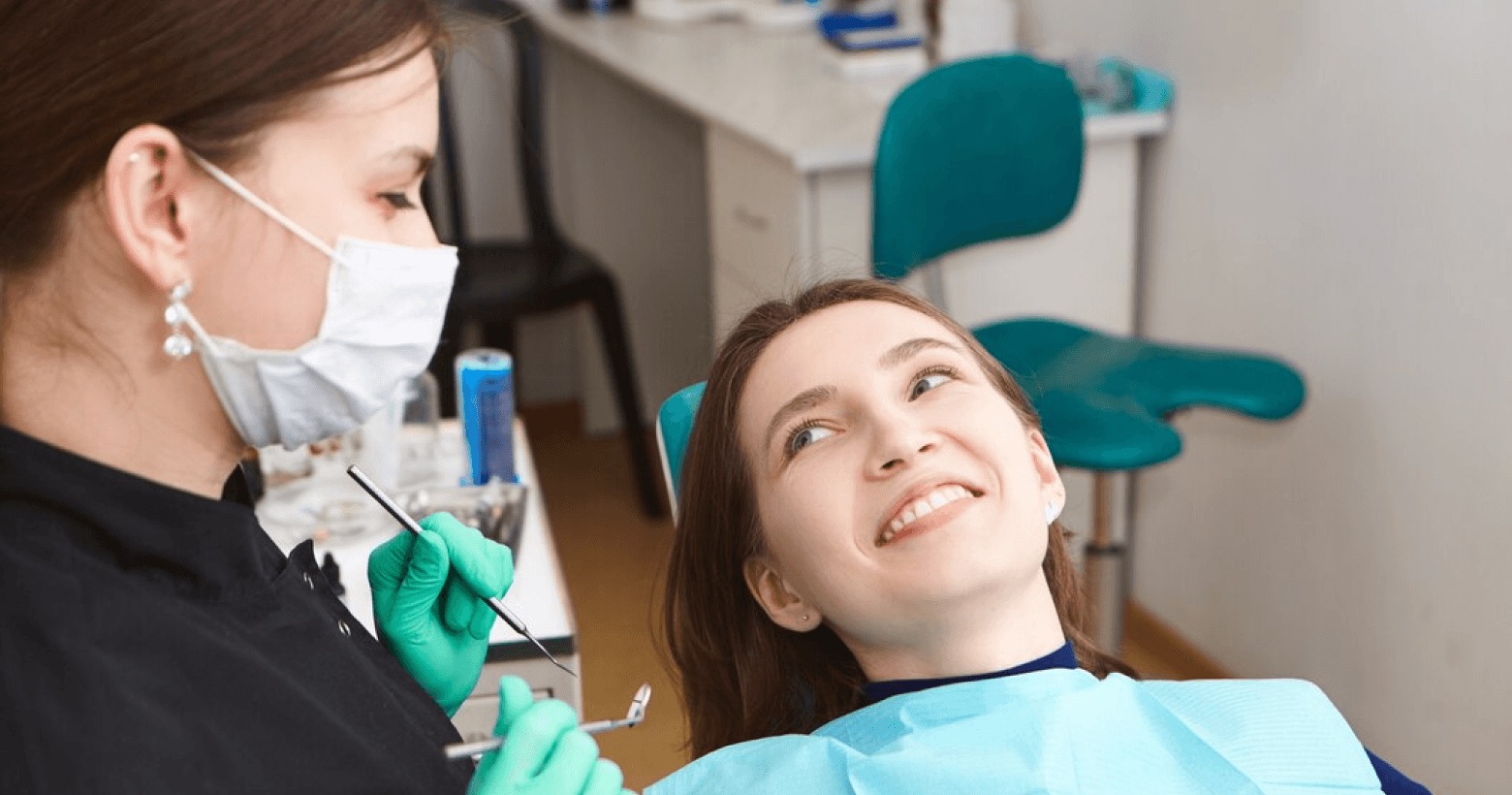 A dental professional wearing a mask and gloves performing a dental procedure on a smiling female patient.