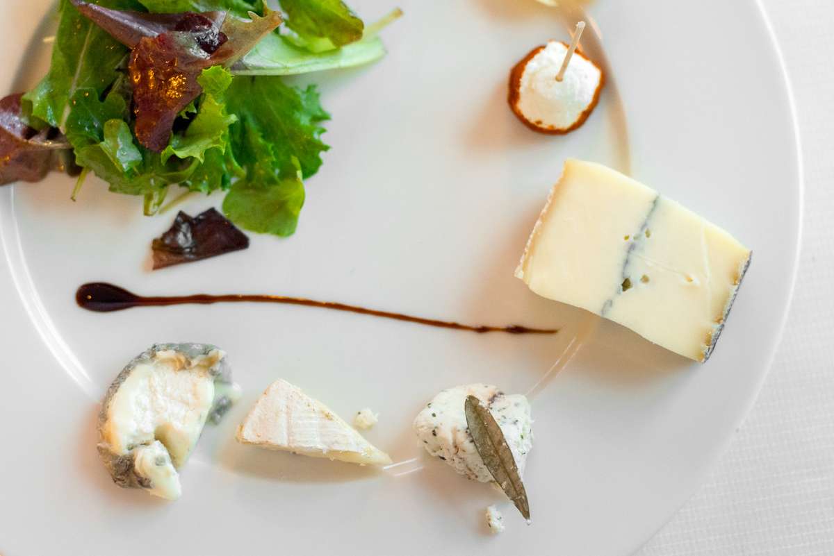 A gourmet cheese plate featuring blue cheese, goat cheese, and a small salad with greens and dark fruit on a white plate.
