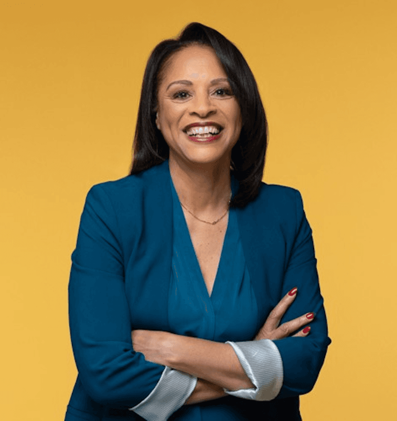 Celeste Warren smiling with folded arms in a teal blazer against a gold background.