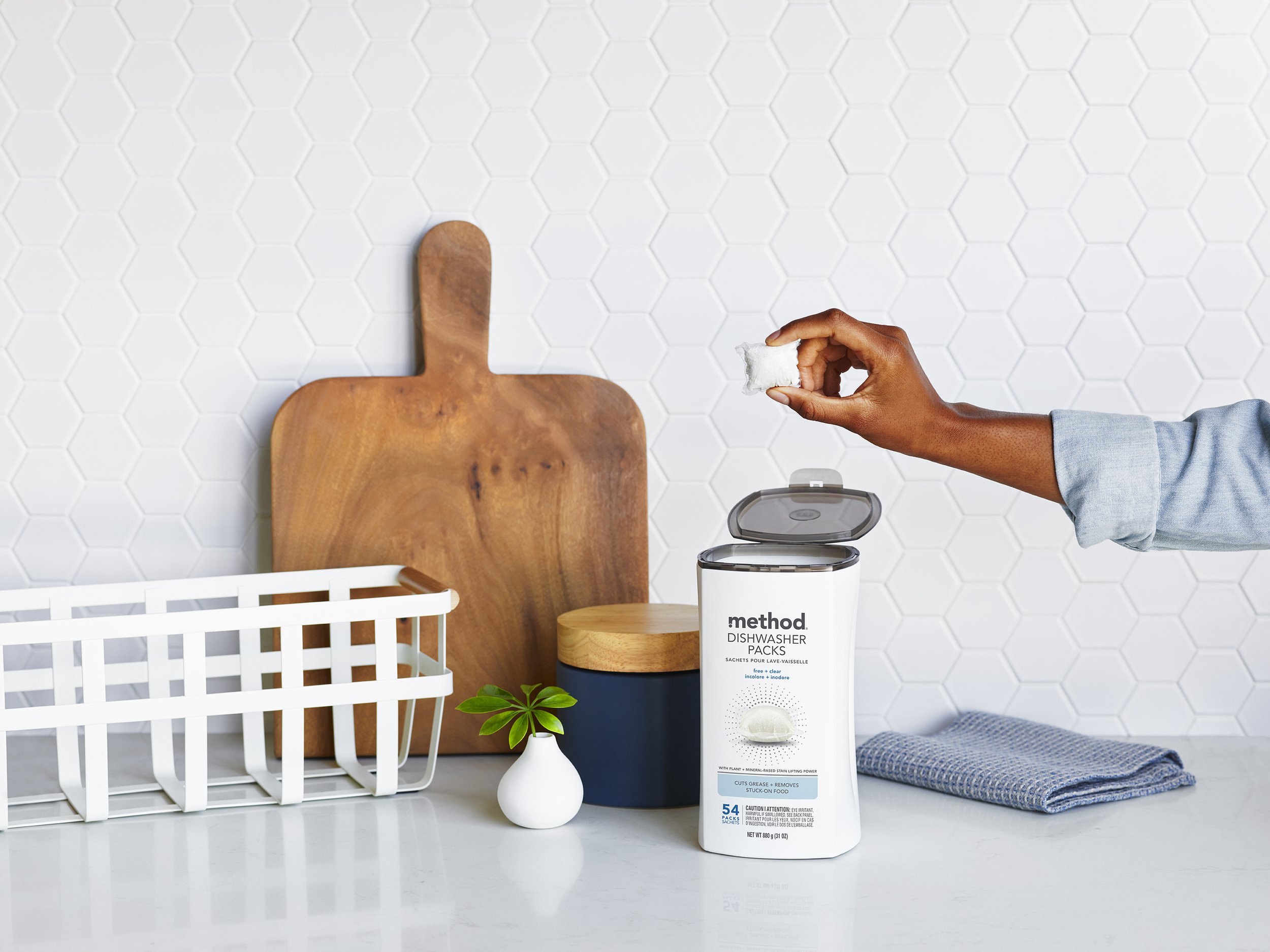 Hand lifting a dishwasher pack out of a white canister with its lid flipped open, on a counter by hex tile
