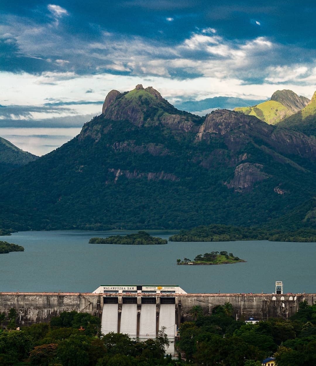 Scenic view of Malampuzha Dam with lush green mountains in the background and a serene reservoir reflecting the sky. The landscape features small islands and rugged peaks, creating a picturesque natural setting