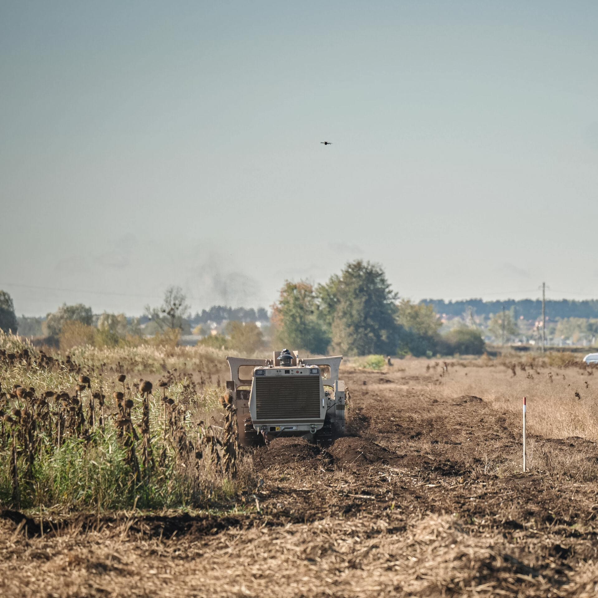 GCS-200 remote-controlled demining machine clearing a minefield in Irpin, Ukraine, with a reconnaissance drone visible overhead.