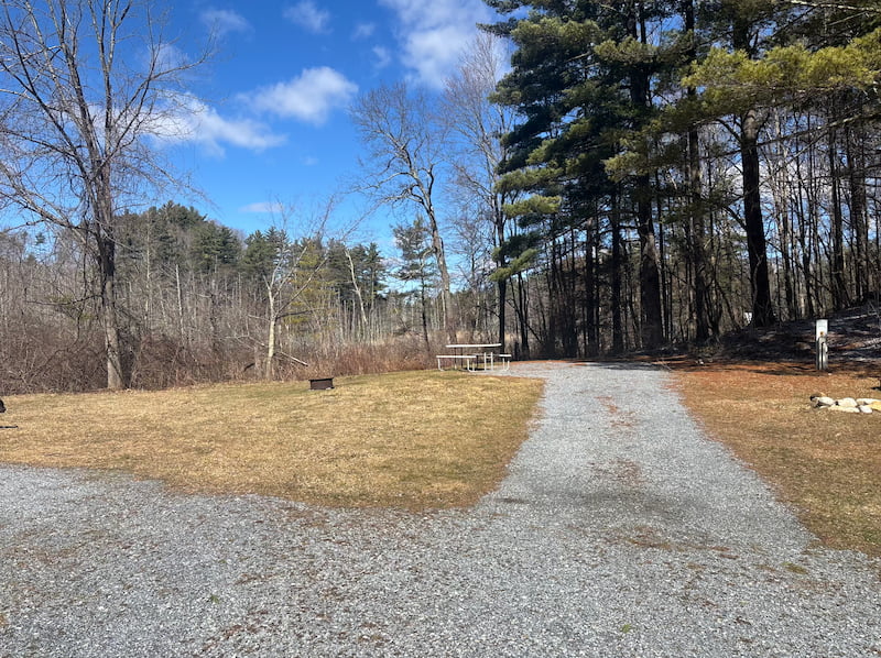 Gravel RV site at Pine Hollow with picnic table, fire ring, and forest view under a blue sky