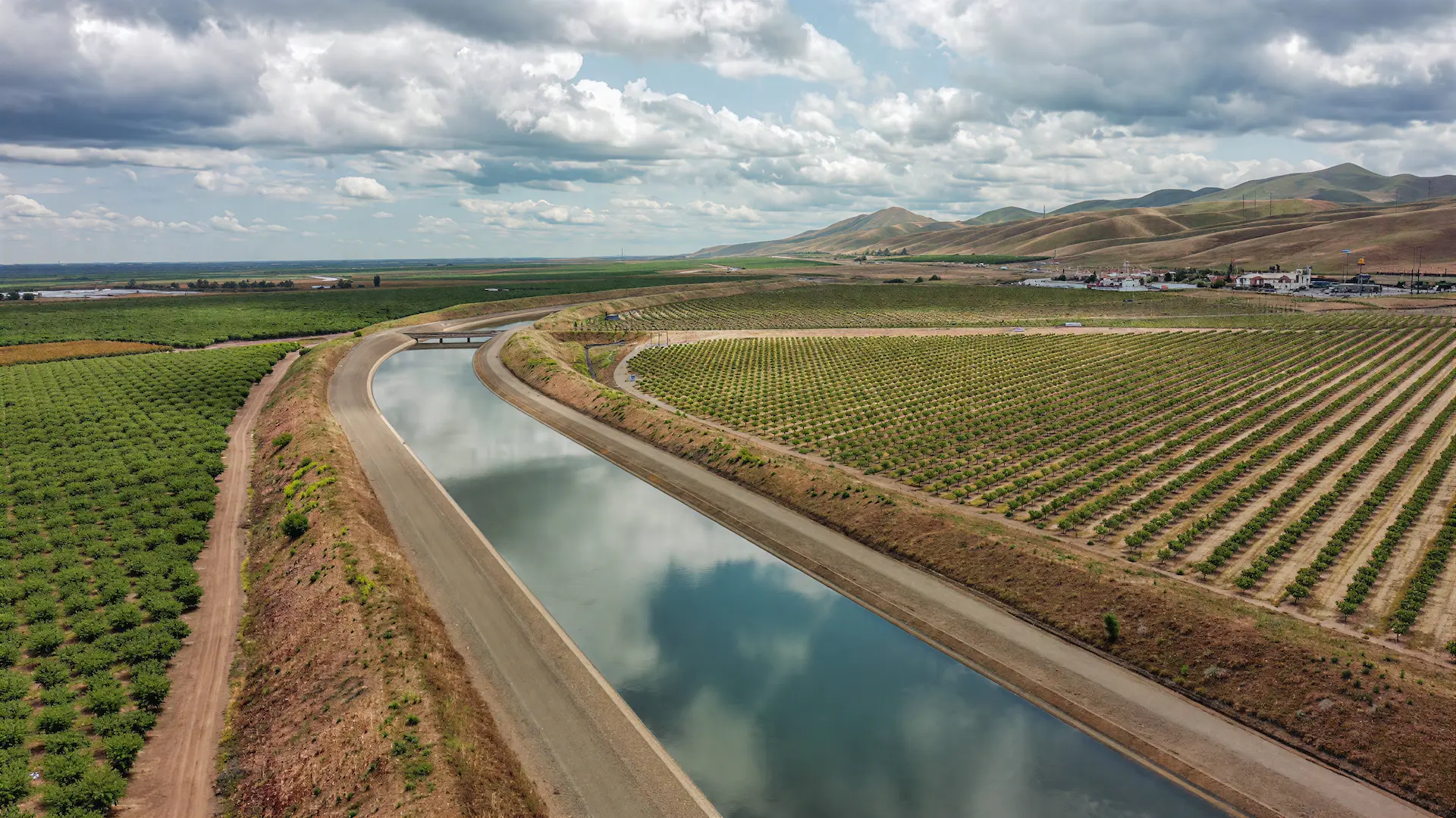 Irrigation canal curving through Central Valley farmland with foothills under dramatic clouds.