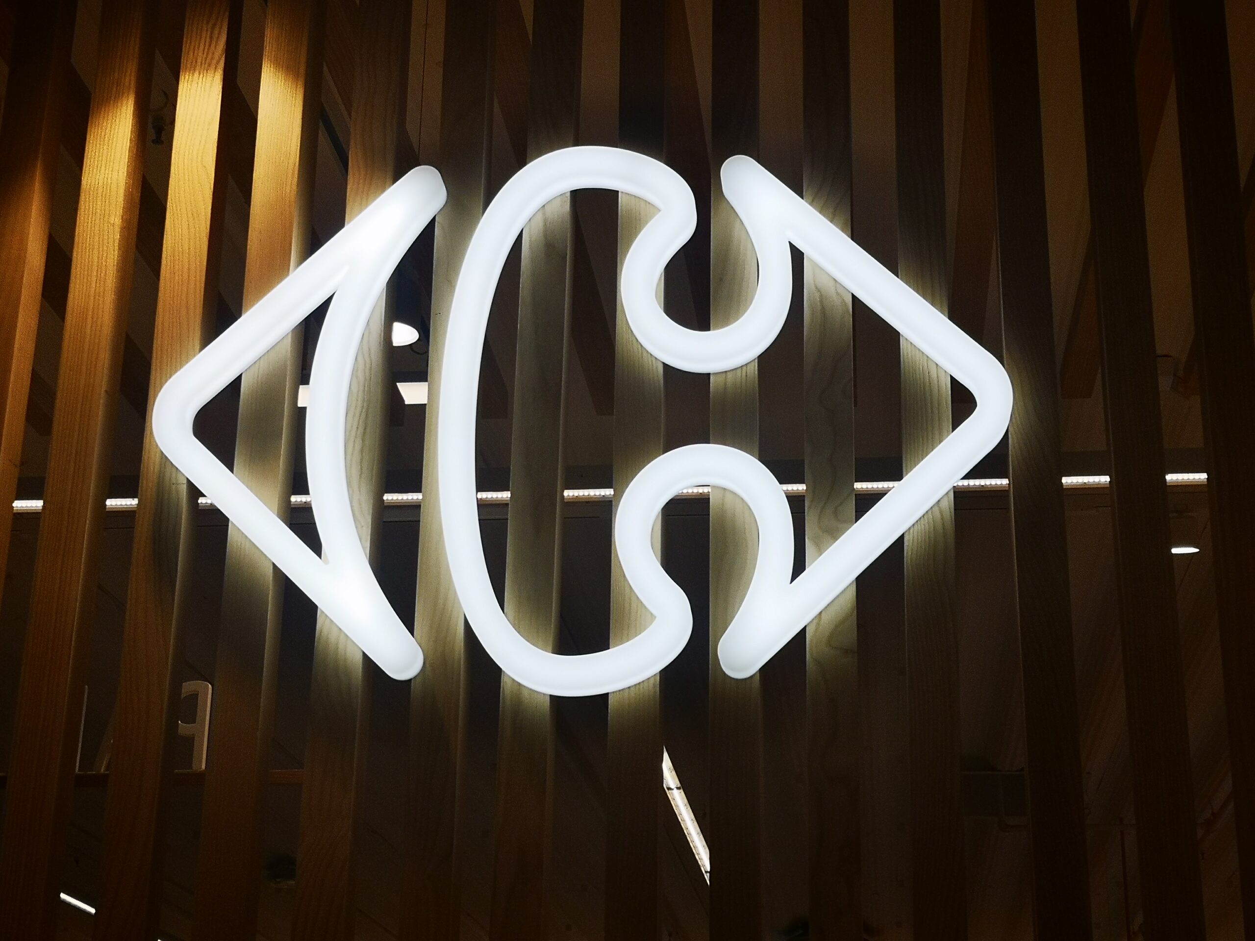 Logo blanc lumineux en forme de flèche sur un fond en lattes de bois. Le design présente des formes courbes symétriques qui lui confèrent un aspect moderne et élégant.