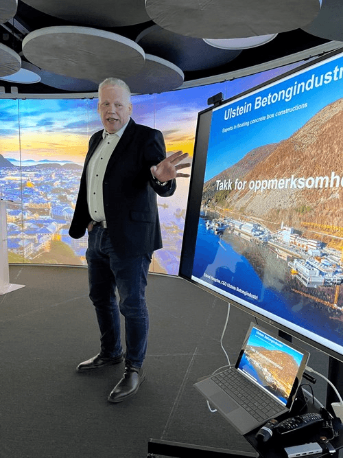A person stands in front of a large screen, gesturing while presenting. The background features a colorful digital display.
