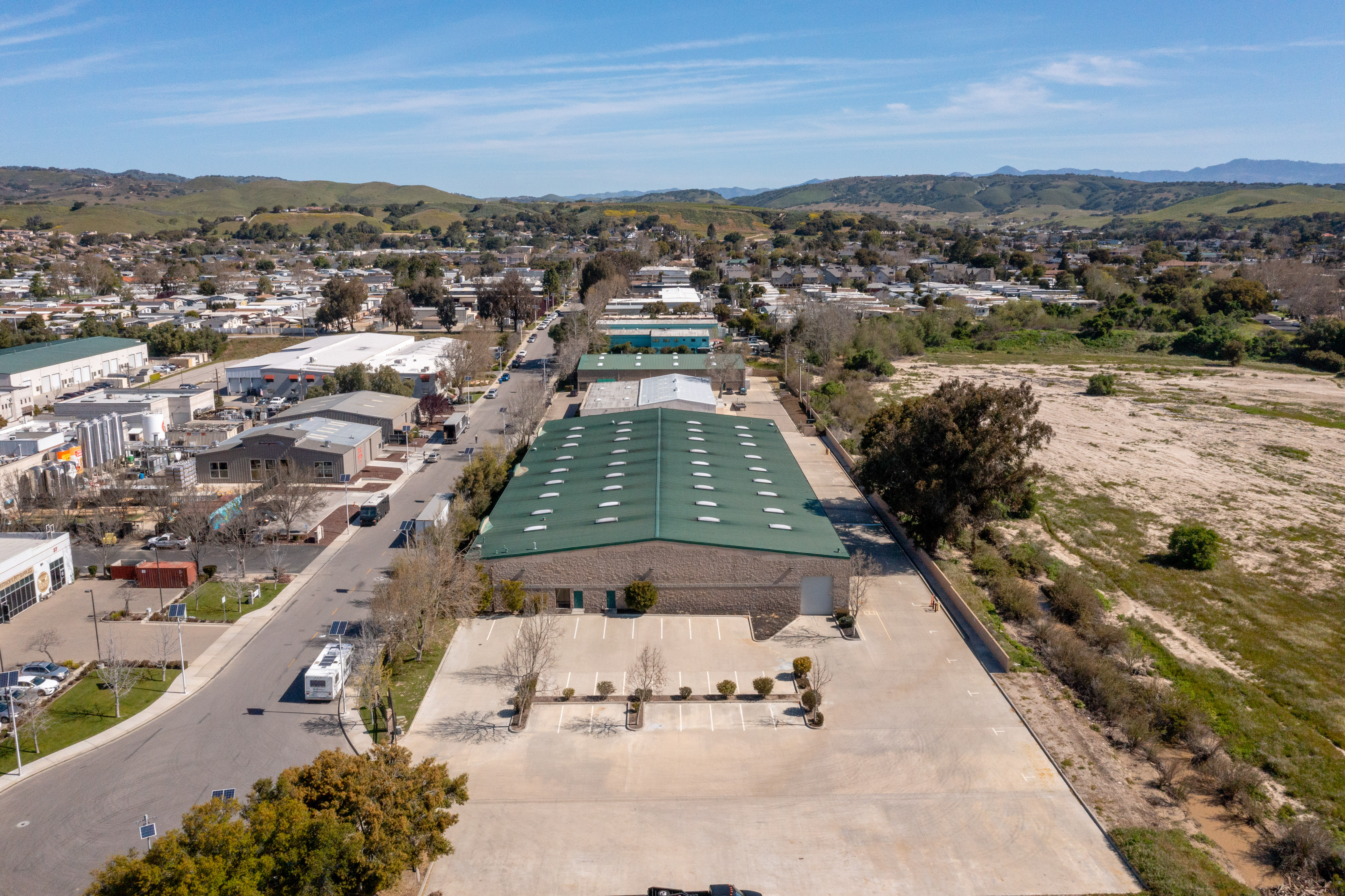 Aerial view of a single-story industrial or self-storage facility with a green standing-seam metal roof and skylights, surrounded by a large paved lot, set within a small California town with rolling hills in the background.