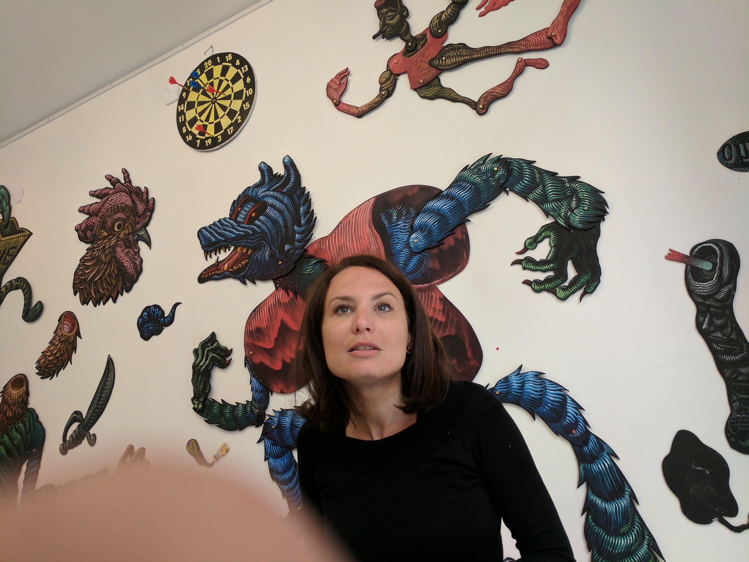 A woman stands before a wall of cut-out painted creatures, a blue wolf-like figure rising behind her.
