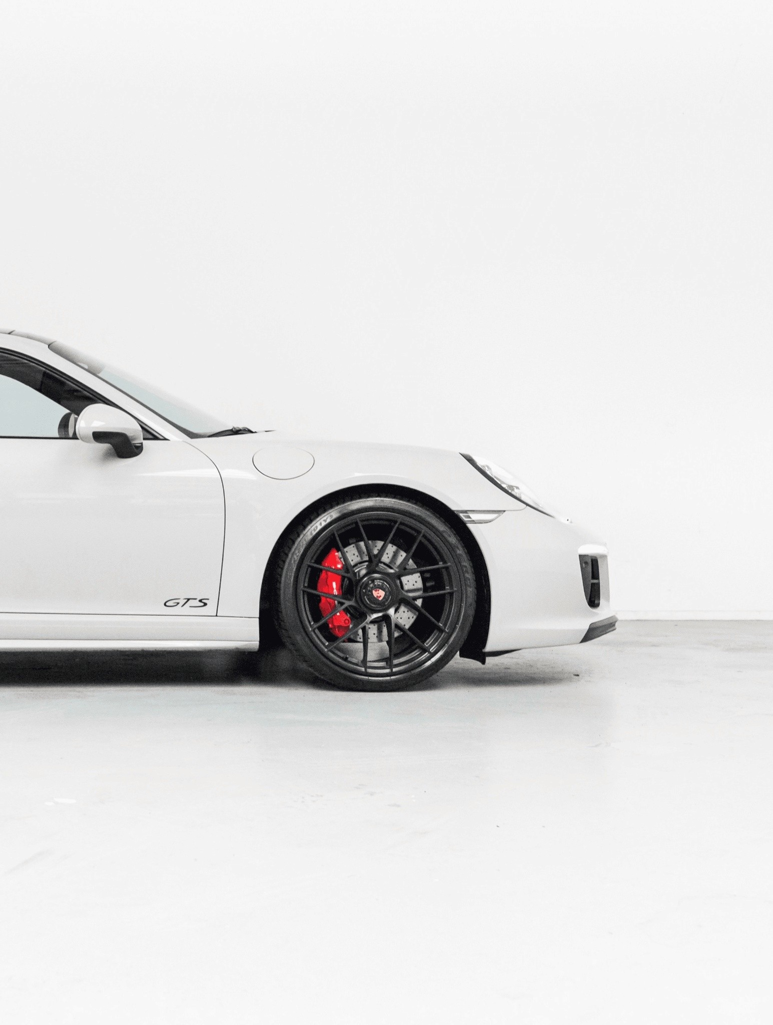 A sleek, white sports car with a distinctive GTS badge sits in a minimalist setting, showcasing its black alloy wheels with red brake calipers against a clean, white background.
