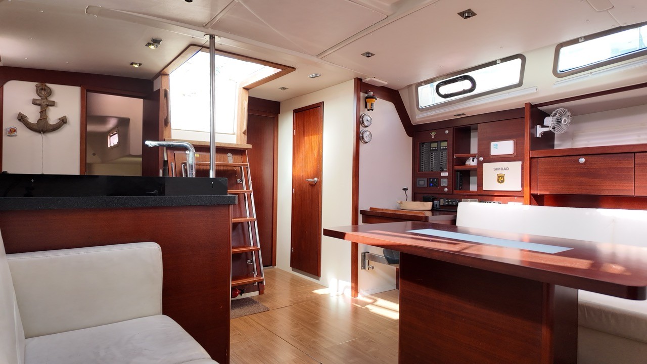 Sailing yacht saloon interior showing EcoDeck light oak composite flooring throughout, with mahogany cabinetry, Simrad nav station, chart table, companionway ladder, and overhead hatches providing natural light