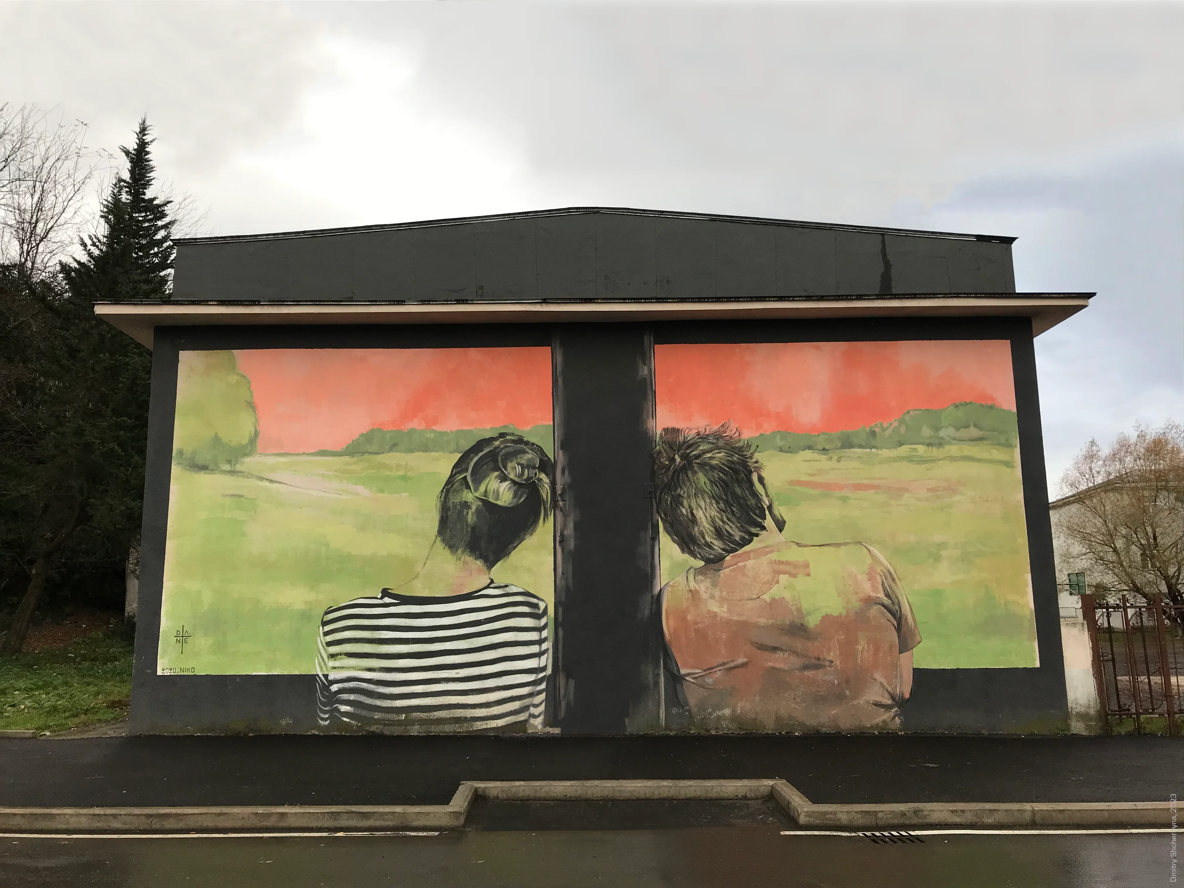 A mural painted on a small building shows the backs of a woman and a man gazing out at a green field. Their views are separated so each see their own picture.