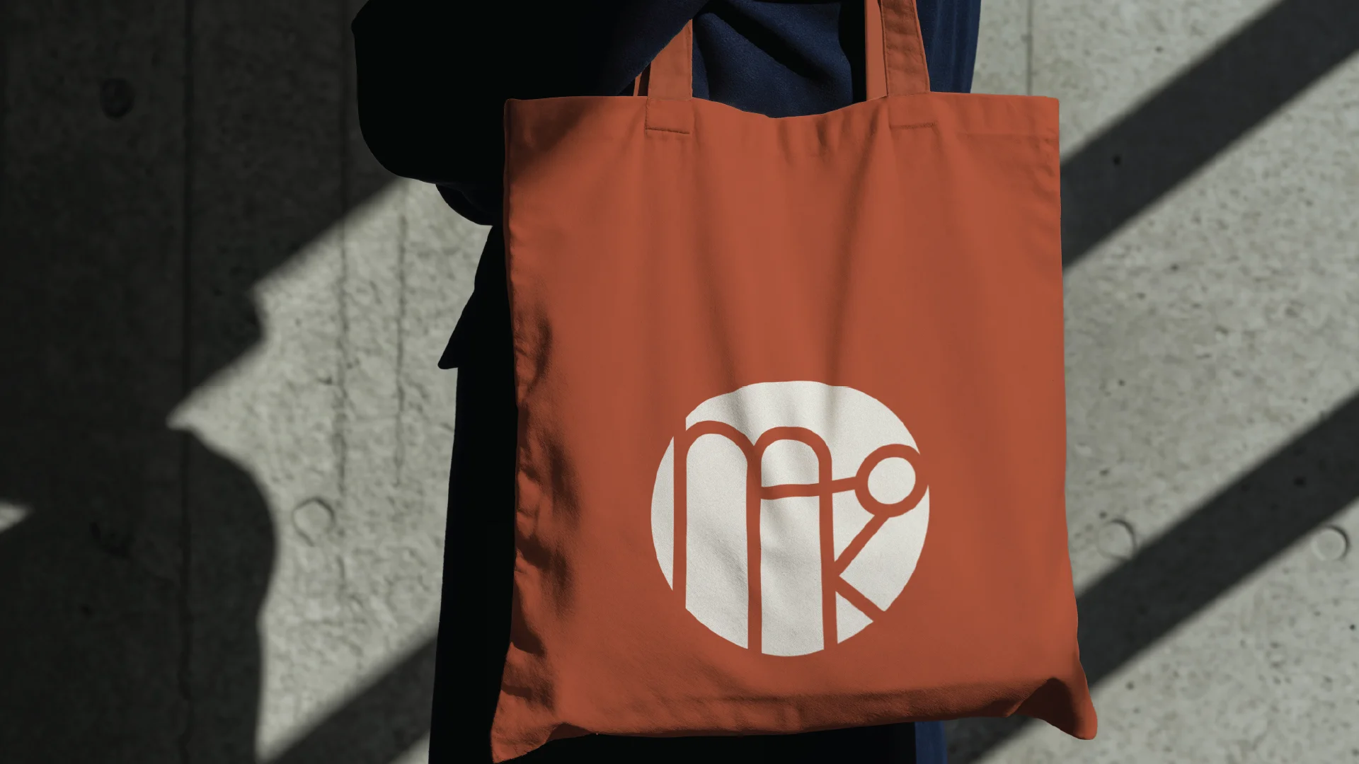 A person holds an orange tote bag with a white circular logo, standing against a textured concrete wall cast with shadows.