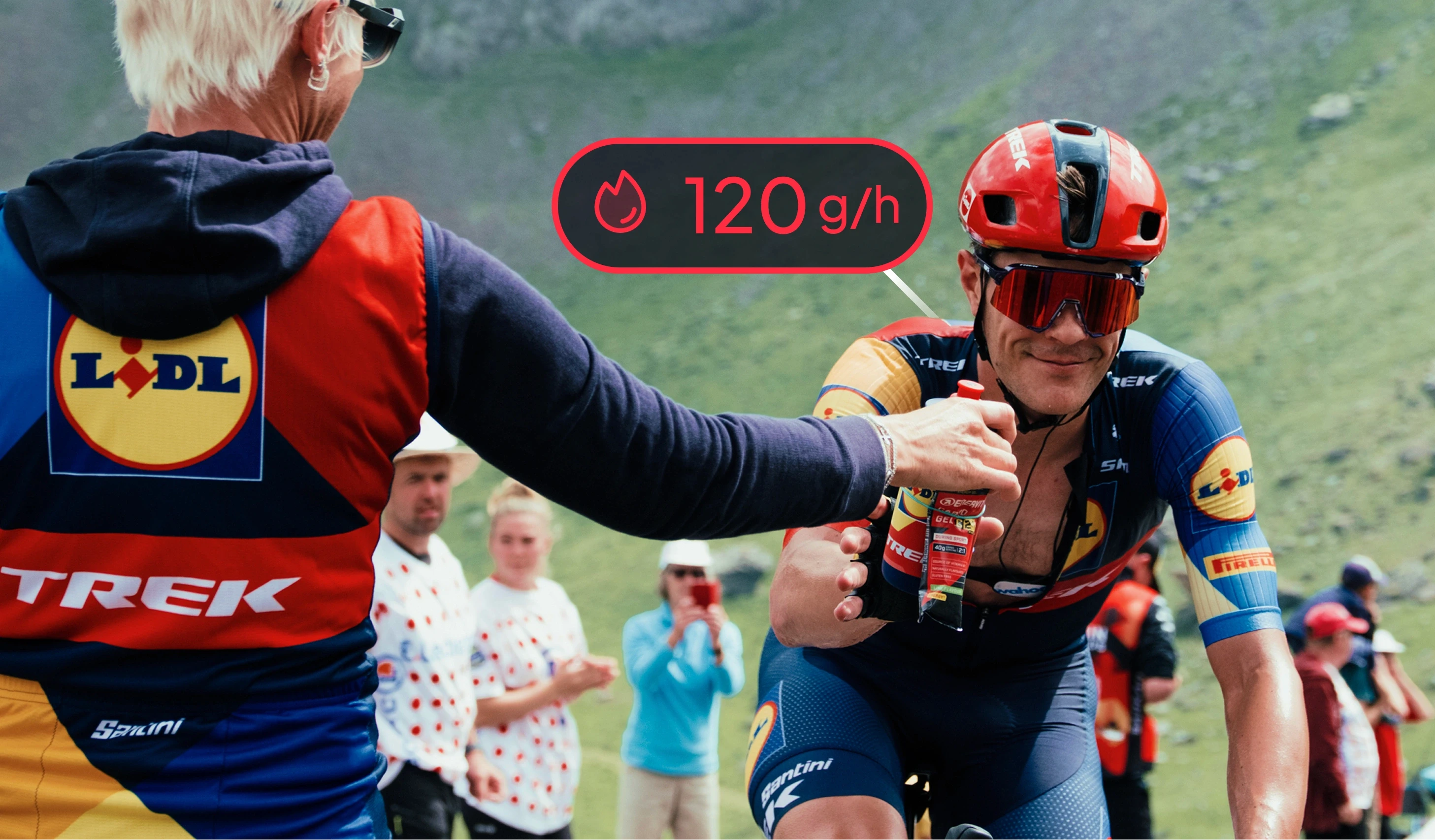 A Lidl-Trek soigneur passes a bidon and a 40g carbohydrate gel to a rider on a mountain climb, with a Vekta carbohydrate target of 120 g/h overlaid