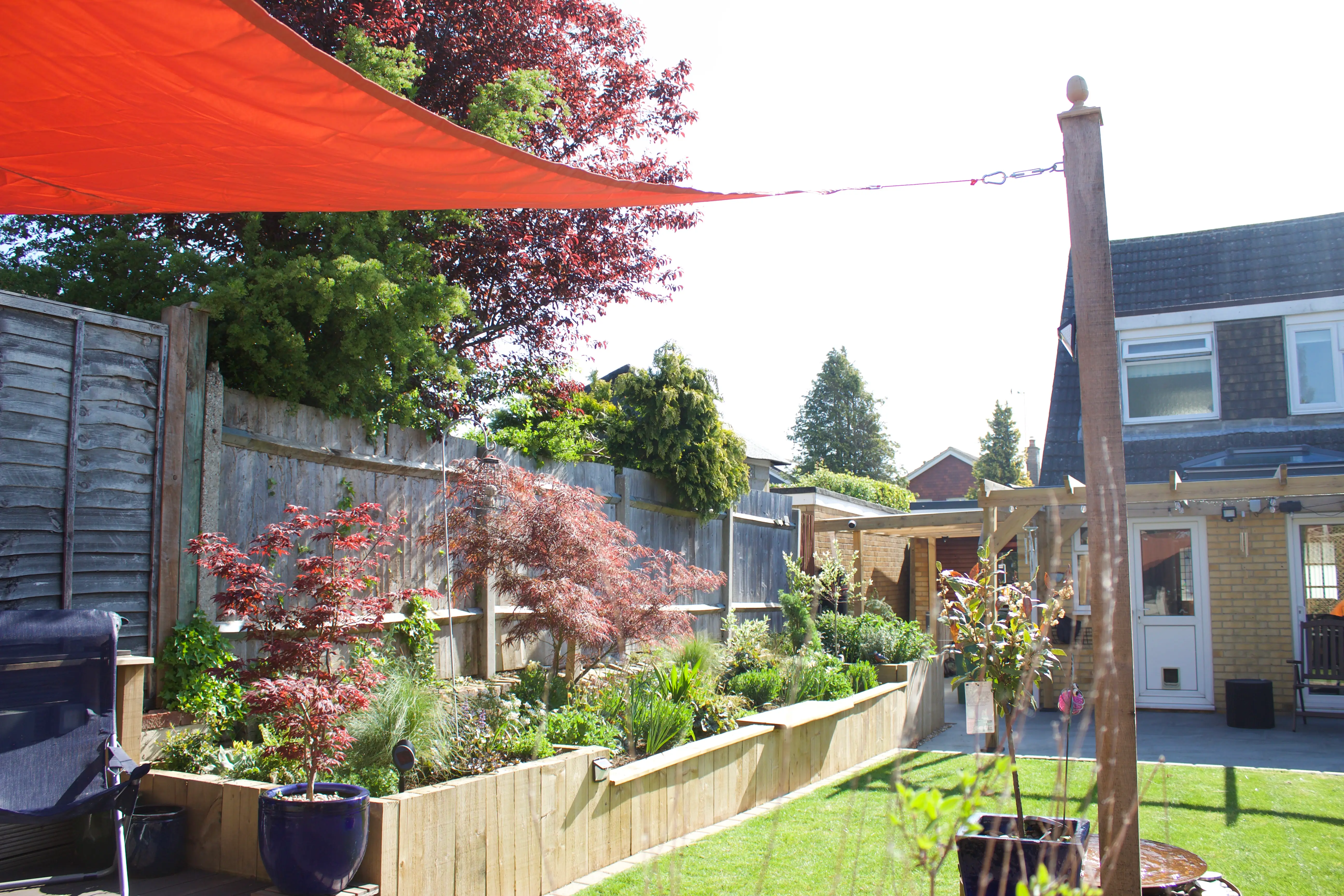 A vibrant backyard garden with colorful plants, a bright orange sunshade, and a cozy home in the background.