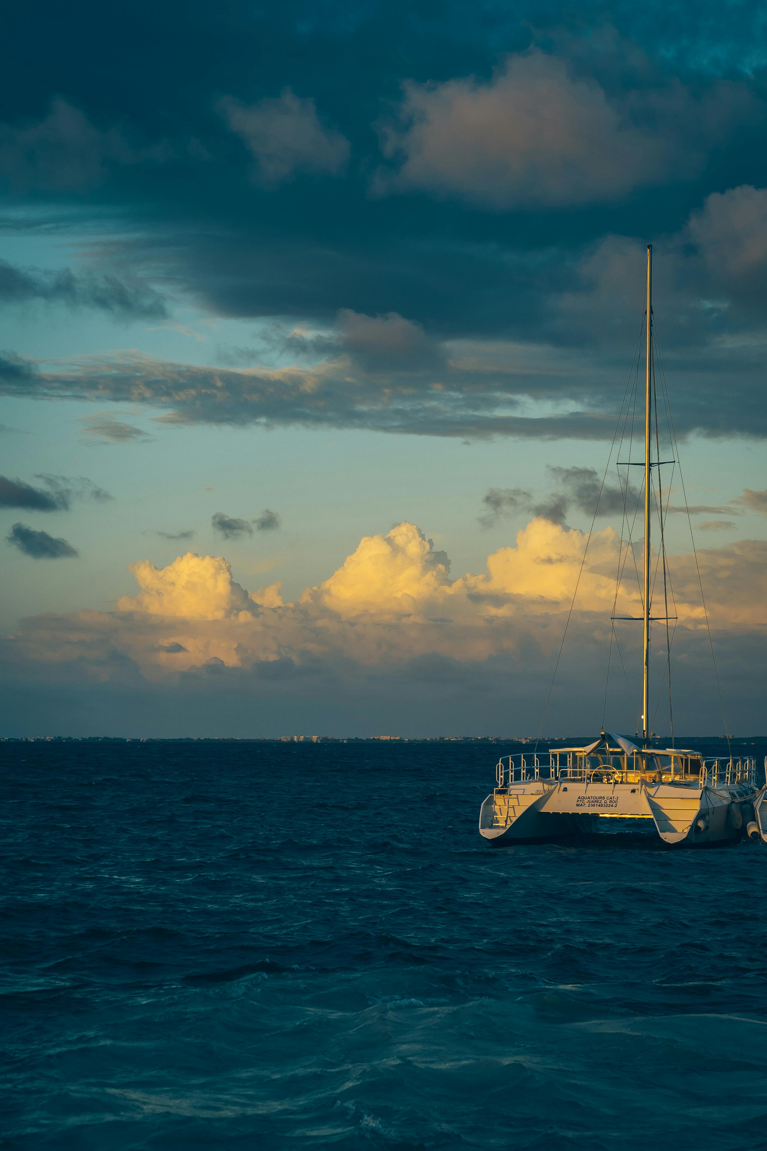 A serene sailboat gently floats on the deep, azure sea under a dramatic sky filled with fluffy, illuminated clouds during sunset.