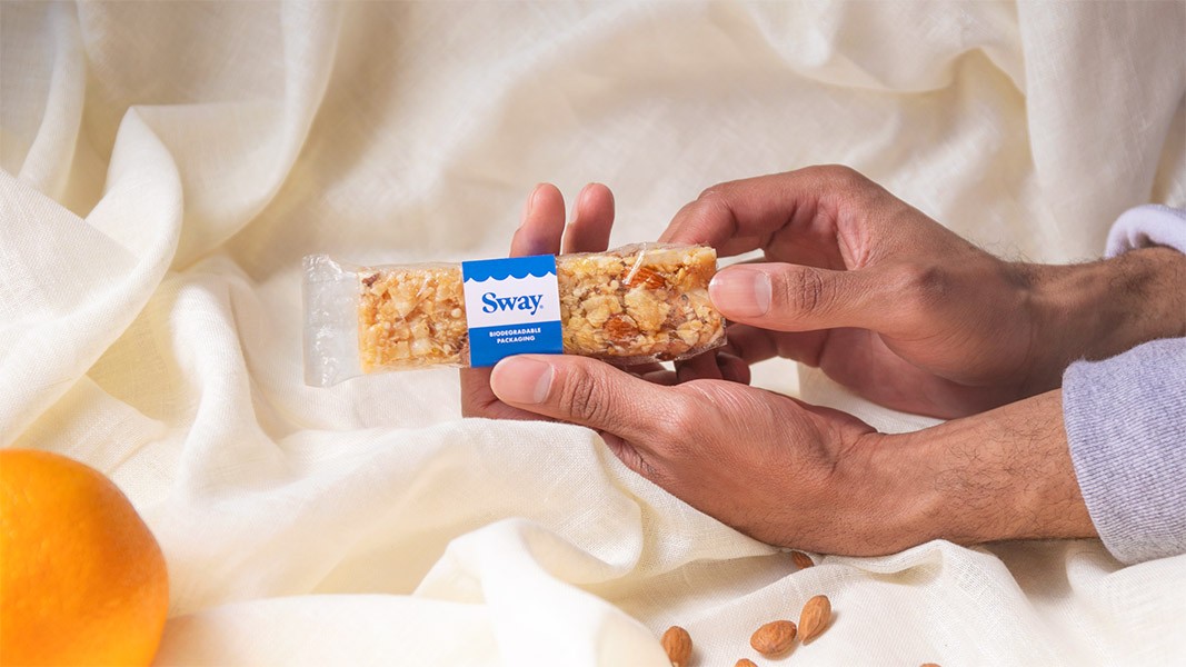 Two hands hold a clear plastic Sway bar wrapper over a white cloth. The wrapper has a blue label reading “Sway” with “Biodegradable Packaging” written below. A partially visible orange and scattered almonds are on the cloth near the wrapper. The hands are positioned over a light-colored fabric surface.