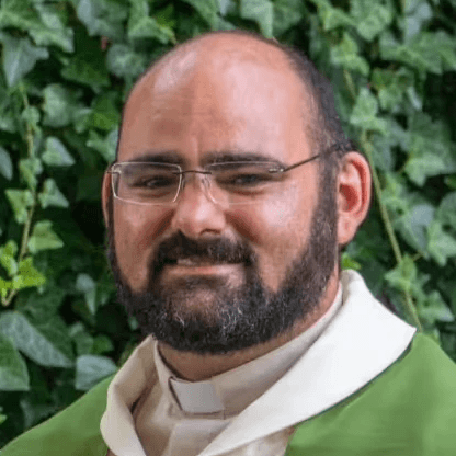 Pfarrer Ludwig Westermeier steht mit Priestergewand vor einer Hecke und lächelt