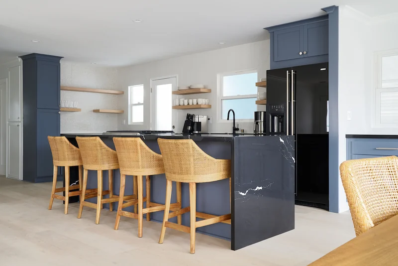 Spacious kitchen interior with navy blue island and matching cabinets, wooden stools, and floating shelves. Natural light enhances the cozy ambiance. Photo by Todd Huge.