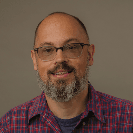 A smiling man with short hair stands with arms crossed, wearing a dark shirt against a neutral background.