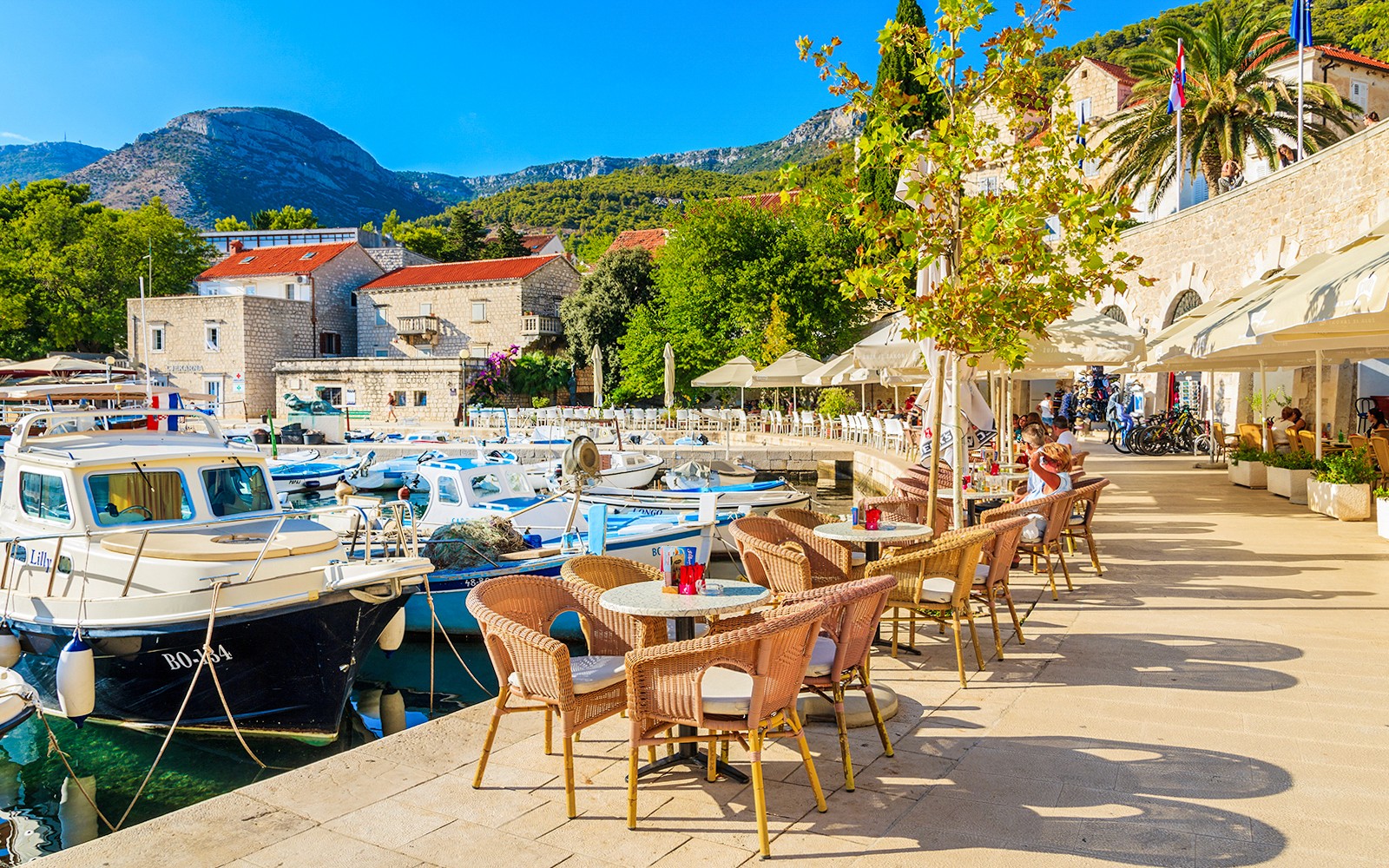 Boats docked at a sunny harbor in Hvar, Croatia, with outdoor seating and scenic mountain views.