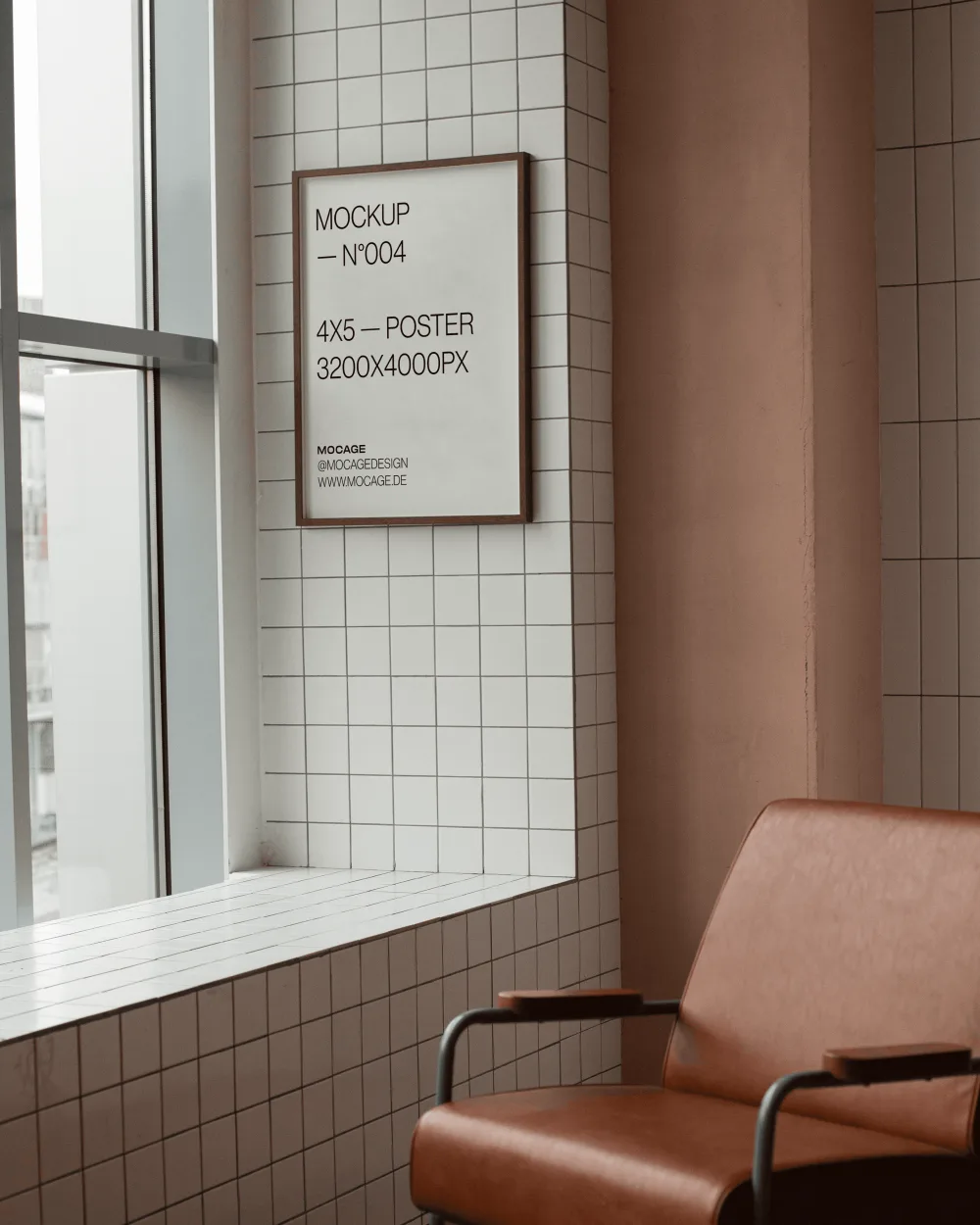 Framed poster mockup on a white tiled wall next to a large window, featuring soft natural light. A brown leather chair with green metal armrests sits in the foreground, adding a modern interior touch.