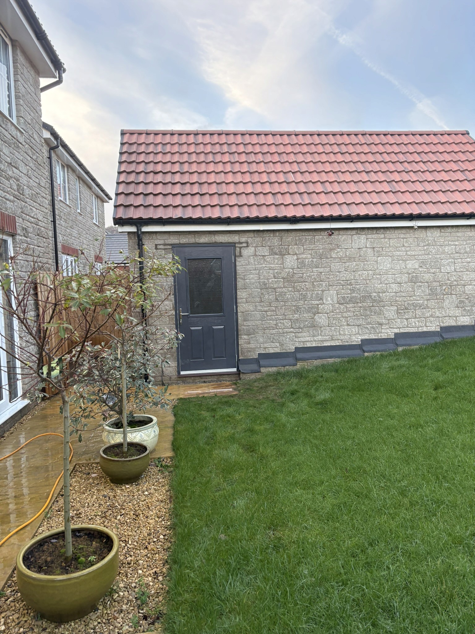 Stone garage with a dark pedestrian access door and red tiled roof, beside a garden lawn and paved path.