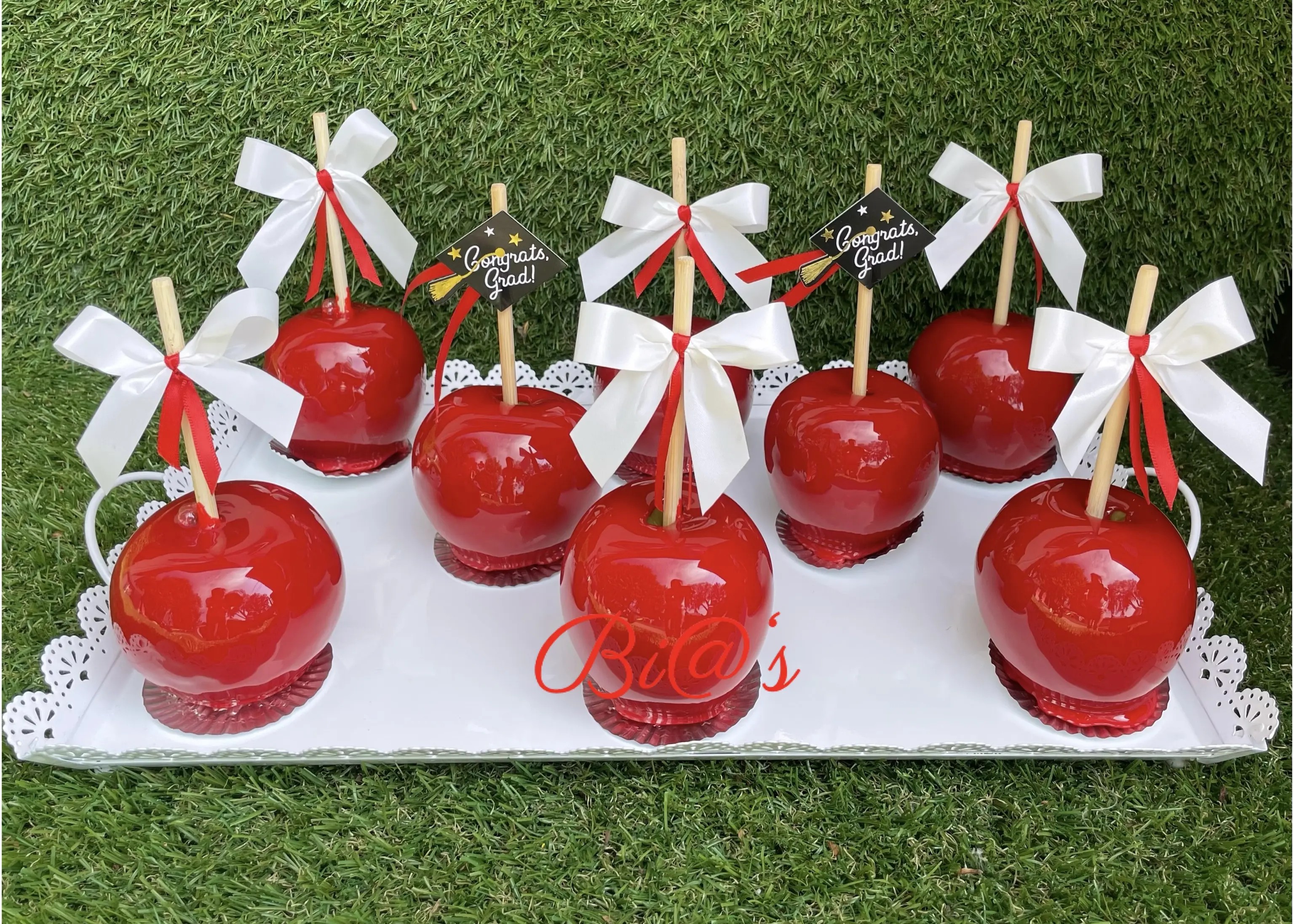 Candy apple display featuring glossy red apples with elegant ribbon ties, arranged on a white tray. A classic treat presented in a refined and festive style. Susa’s Sweets & Balloons.