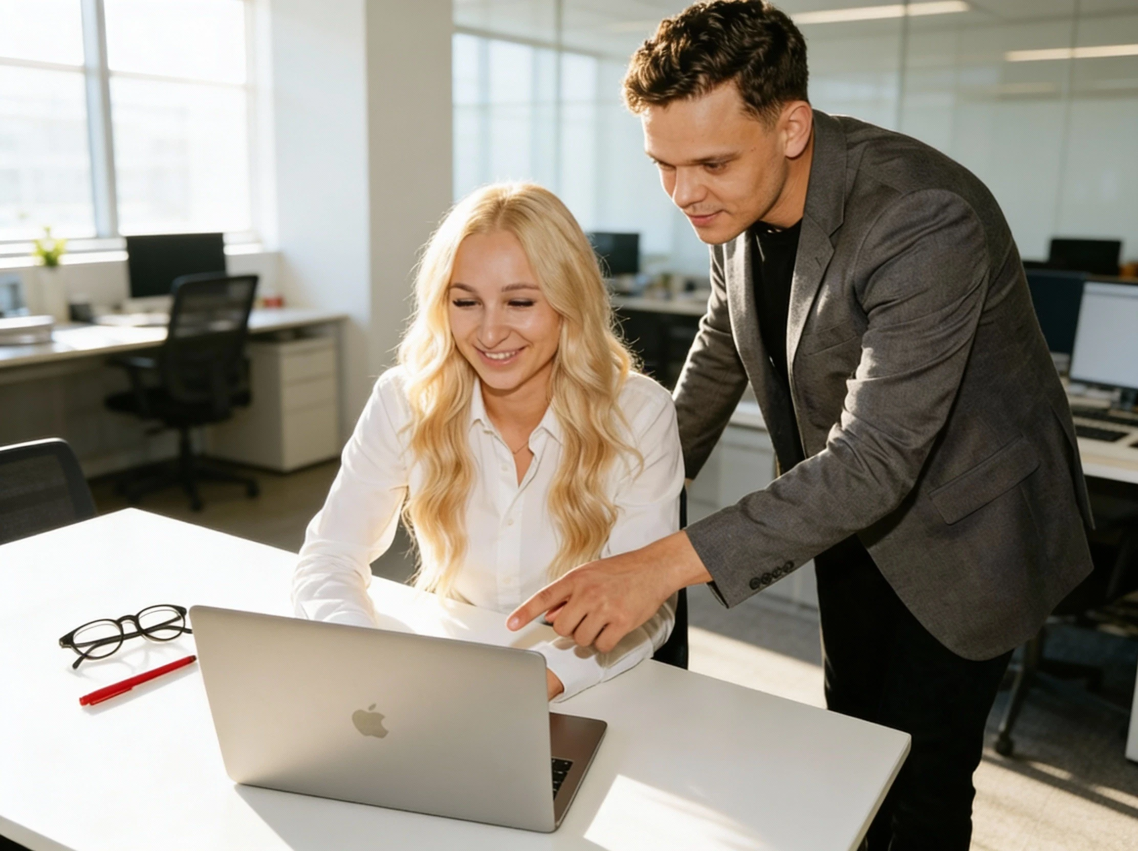 A man in a dark sweater and a woman in glasses wearing a navy blouse sit together at a desk, smiling at the camera with a laptop in front of them.