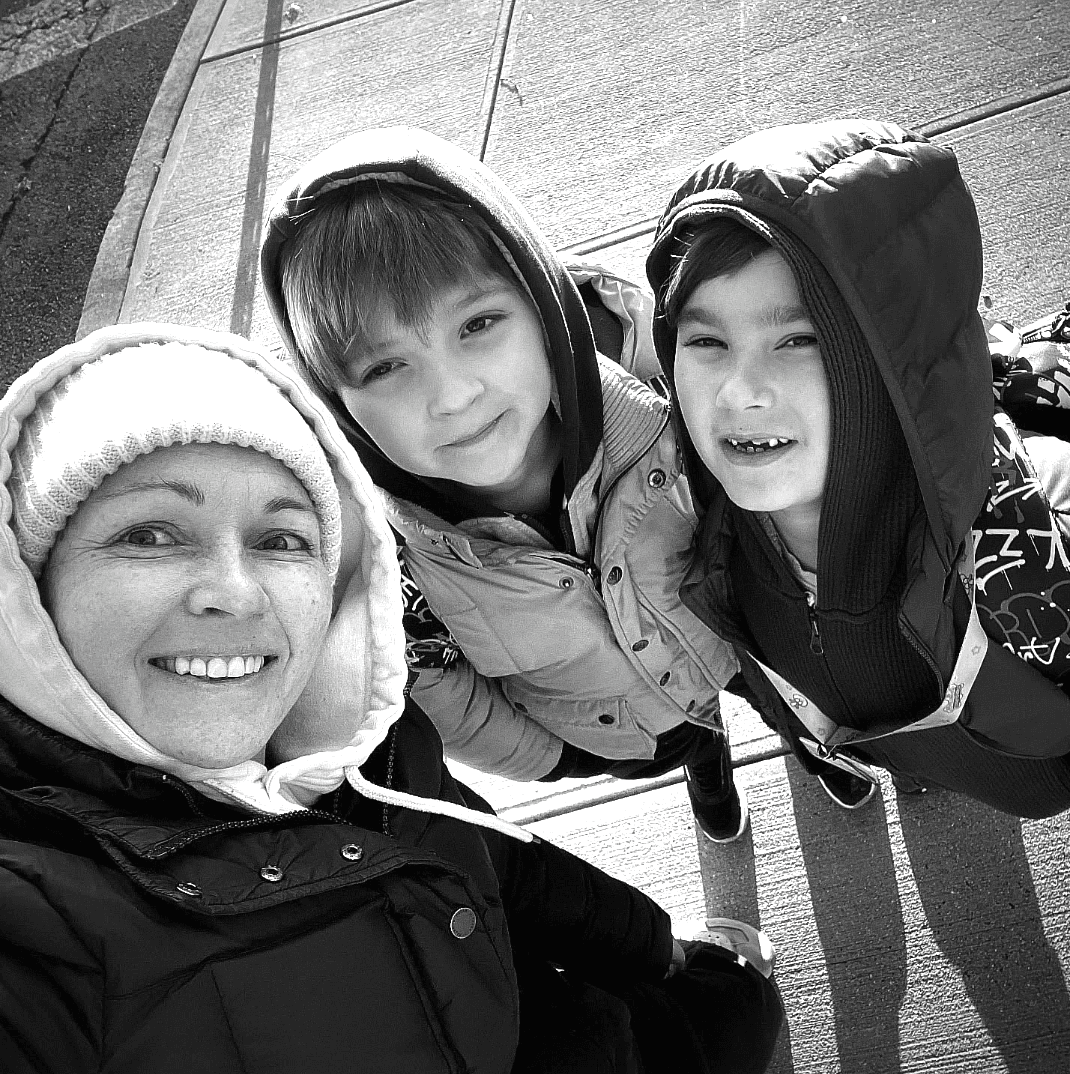 Black and white photo of a mother wearing a hat taking a selfie from above with her fraternal 7.5-year-old twin boys. The boys are wearing warm hoodies and are looking up towards the camera with a sense of wonder and affection.