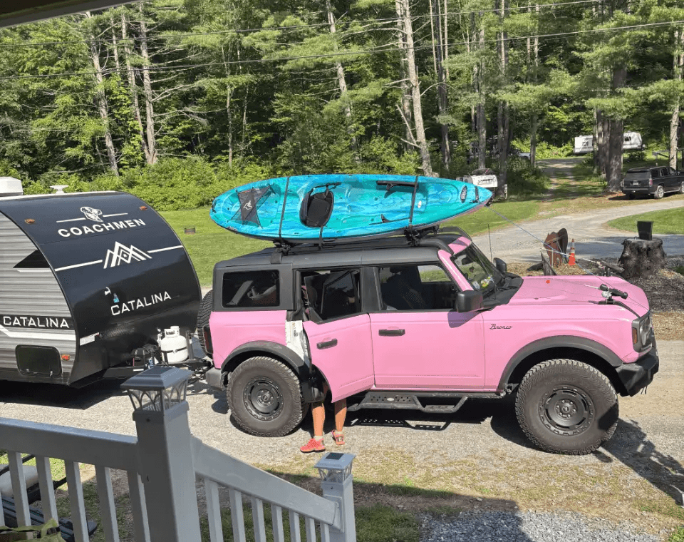A bright pink Ford Bronco towing a Coachmen Catalina trailer with a kayak on top, parked at a scenic RV site in Pine Hollow Campground.