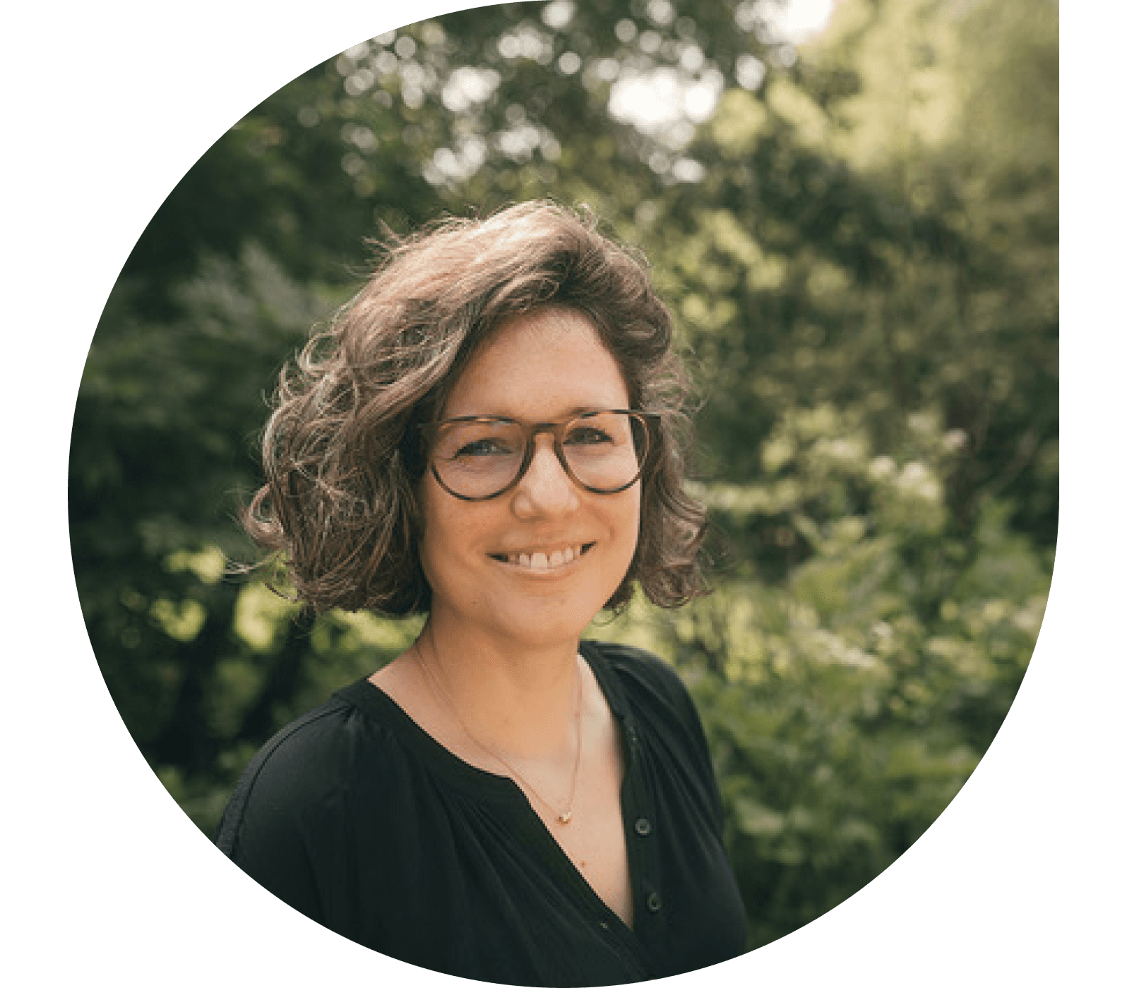 A smiling European woman with glasses, stands in a sunny park. She has a warm, approachable demeanor, with wavy hair and a professional yet casual outfit