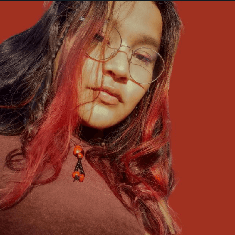 A woman with long dark hair with red highlights and glasses, posing against a simple reddish background.