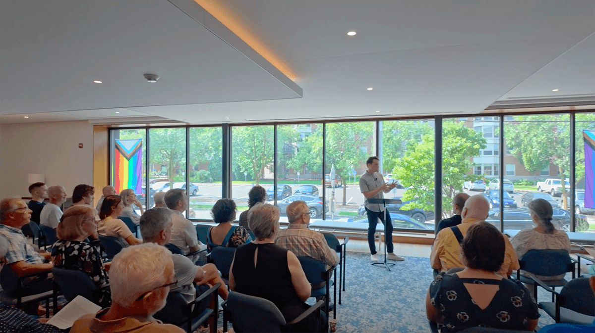 Grace Trinity Church congregation gathered for a presentation in a bright room with large windows, a speaker at the front, and Pride flags displayed in the background.